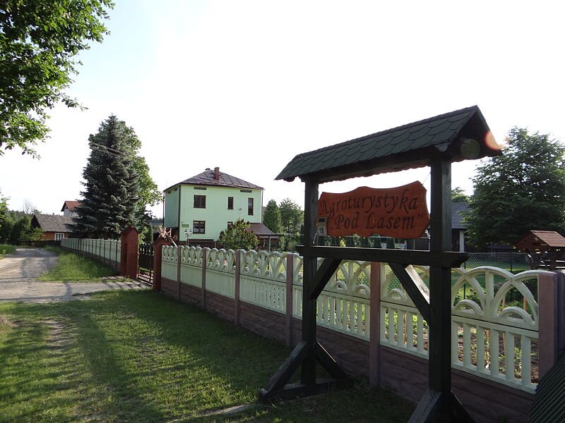 Fotografia przedstawia gospodarstwo agroturystyczne w Szwedach na Podkarpaciu. Gospodarstwo otoczone jest betonowym płotem, na podwórku znajduje się wysoki zielony budynek. Otaczają go drzewa iglaste. Na pierwszym planie wyróżnia się drewniany szyld z napisem: Agroturystyka „Pod lasem”. 