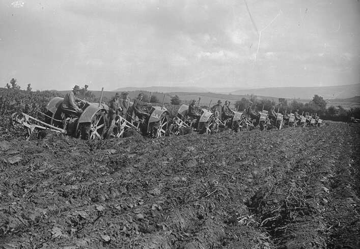 Zdjęcie przedstawia zaorane pole oraz ustawione w rzędzie traktory. Na kierownicą widzimy siedzących mężczyzn. Obok stoją inni mężczyźni. Wszyscy zwróceni są w kierunku kadru, pozując do zdjęcia. 