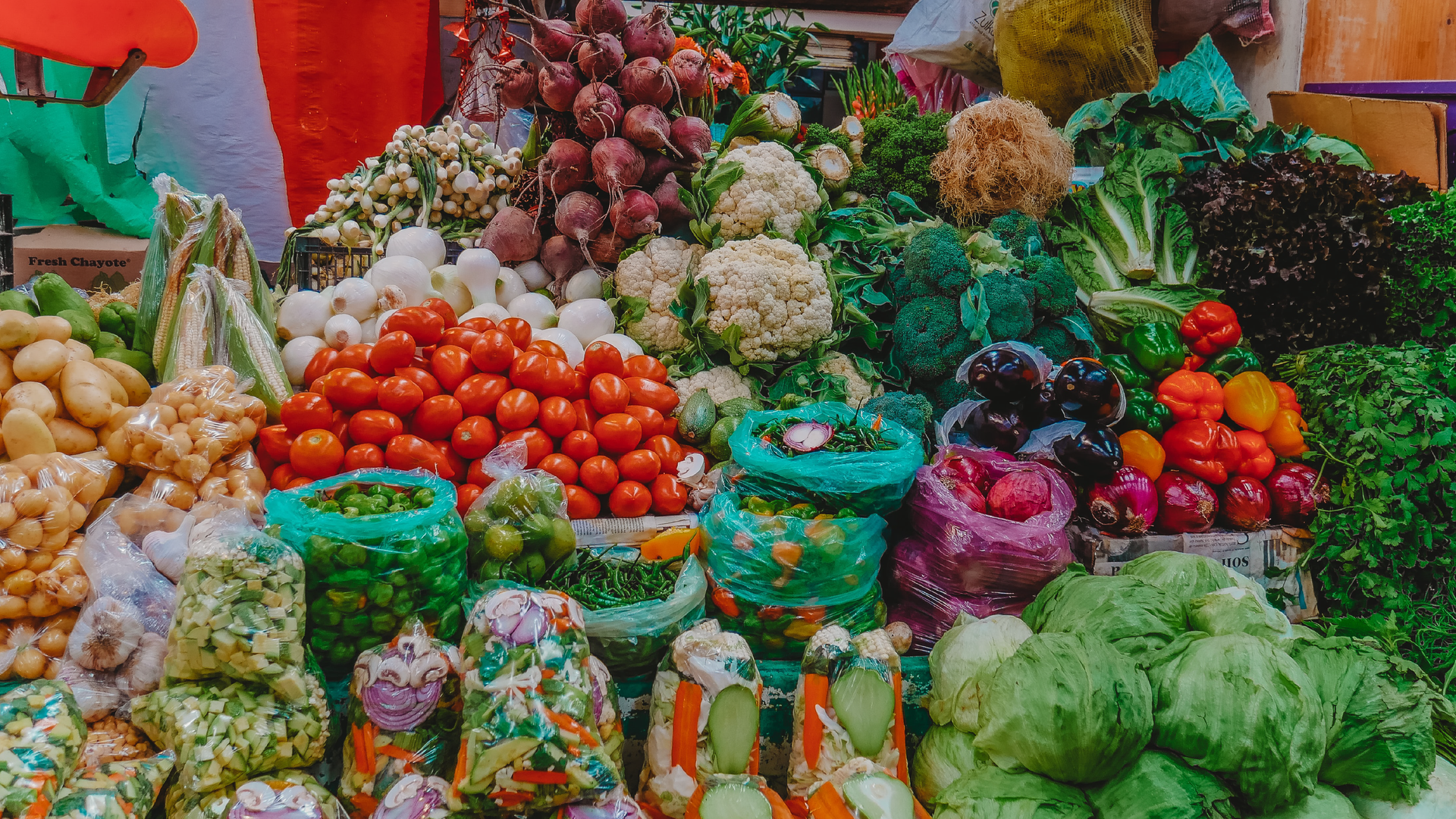 Zdjęcie przedstawia stragan z dużą ilością warzyw. Na zdjęciu znajdują się  poukładane warzywa: ziemniaki, kukurydza, papryczki, cebula, buraki pomidory, kalafiory, brokuły, różne sałaty, różnokolorowe papryki, bakłażany, kapusta i natki.