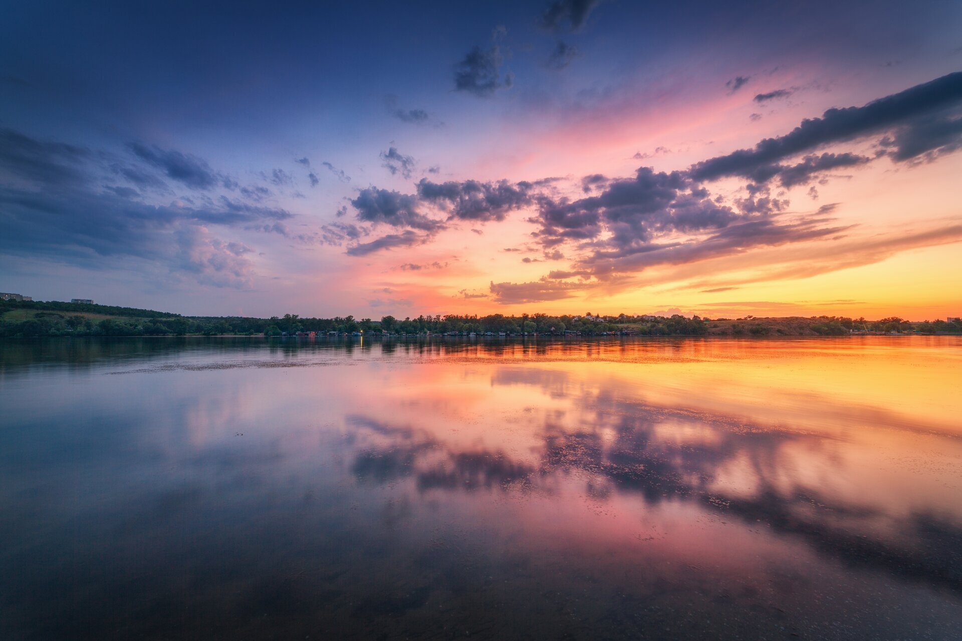 Fotografia przedstawia wschód lub zachód słońca nad jeziorem. Przez środek fotografii ciągnie się ląd. Rosną na nim drzewa. Niebo odbija się w wodzie, sprawiając wrażenie, jakby po obu stronach lądu były chmury. Chmur jest niewiele, są jakby rozciągnięte po niebie. Niebo na linii lądu ma kolor czerwony, im wyżej przechodzi w pomarańczowy, żółty, różowy, jasnoniebieski i ciemnoniebieski. 