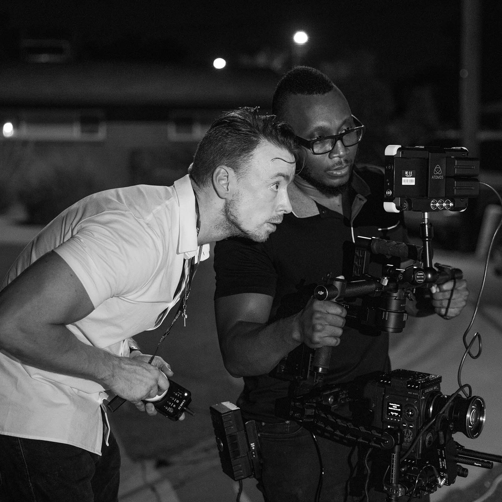 Czaro‑białe zdjęcie ukazuje dwóch mężczyzn stojących obok siebie. Jeden z nich ma zamontowaną kamerę na biodrach, trzymając w rękach rozkładany statyw na którym umieszczono mały ekran. Mężczyzna stojący obok zaglądając przez ramię operatora kamery patrzy w ekran na statywie.