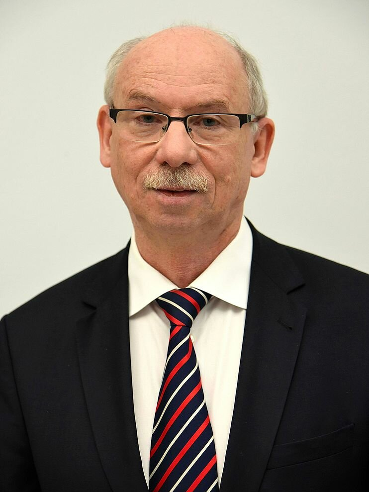 Fotografia przedstawiająca dojrzałego mężczyznę. Postać siedzi, ukazana jest od piersi w górę na wprost nas. Mężczyzna ma owalną twarz, znaczną łysinę, zmarszczone czoło, delikatnie uniesione brwi. Ma nieco opadające powieki, lekką opuchliznę pod oczami z i niewielkie zmarszczki wokół oczu. Mężczyzna nosi okulary, ma duży nos i gęste wąsy. Jest ubrany w koszulę i marynarkę. Na szyi ma zawiązany krawat.