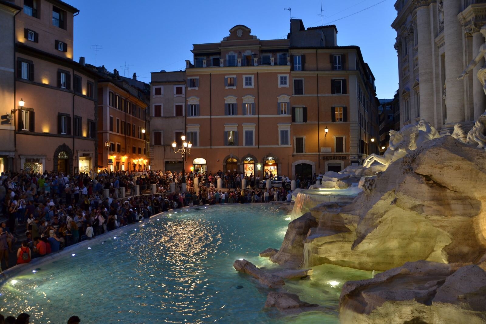 Zdjęcie ukazuje plac w Rzymie ze sławną Fontanną Di Trevi. Dookoła fontanny zgromadził się tłum turystów. Plac otaczają wysokie, stare kamienice, oświetlone przez palące się uliczne lampy. Jest wczesny wieczór, światła odbijają się w wodzie fontanny.