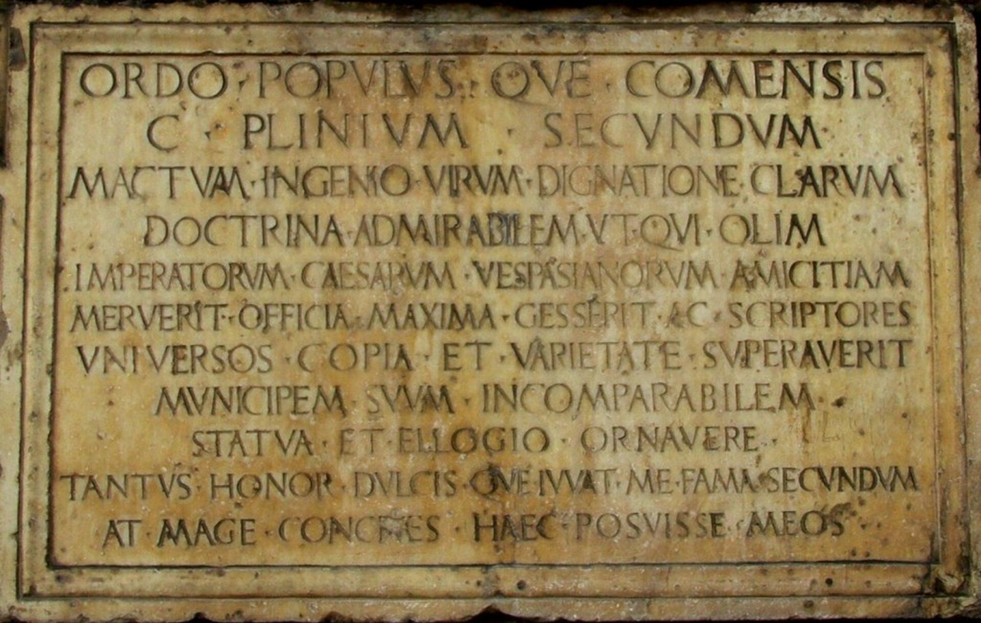 Ilustracja przedstawia łacińską inskrypcję z Como upamiętniającą życie i działalność Pliniusza. Tablica jest w kolorze brązowym. 