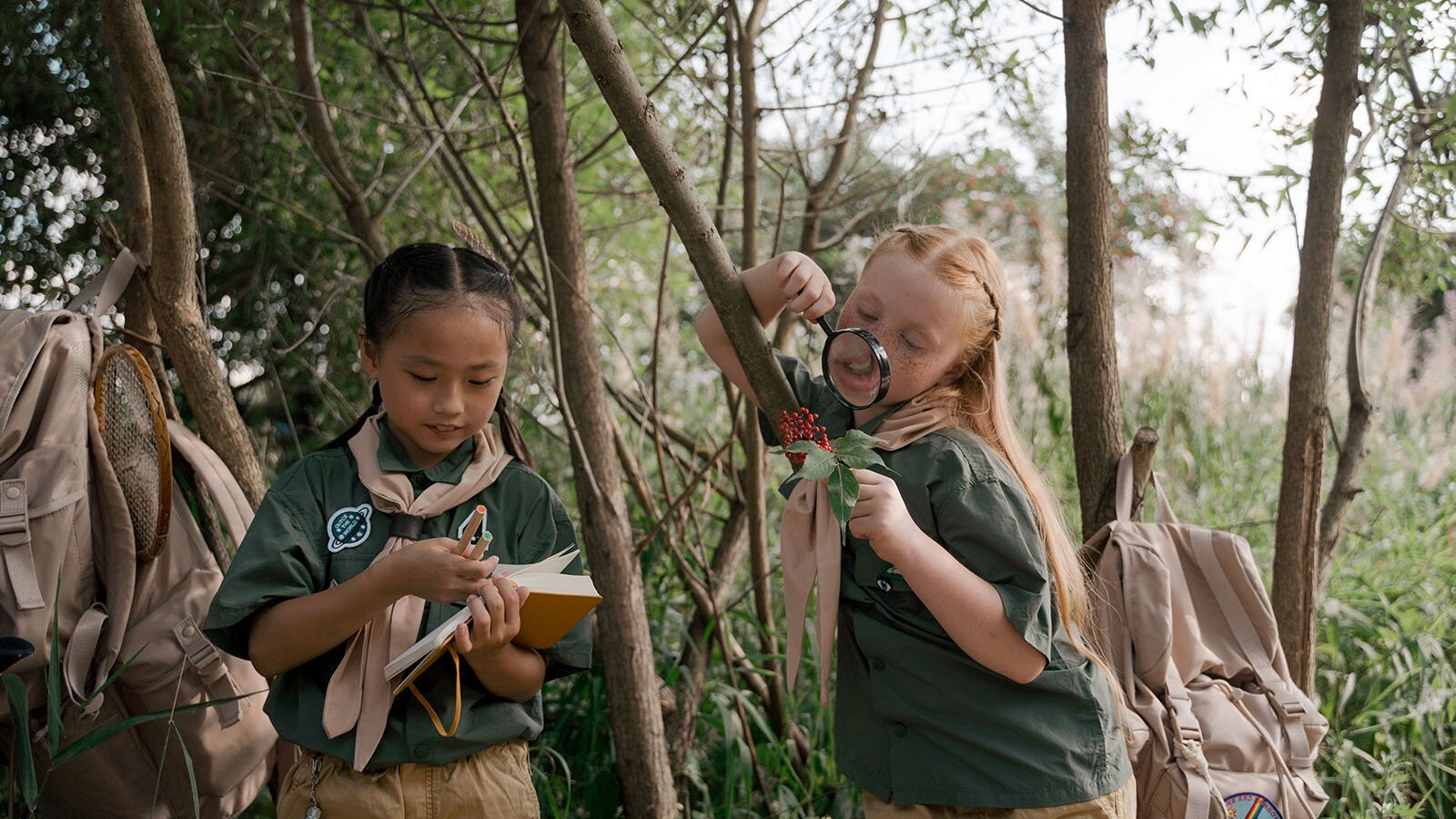 Zdjęcie przedstawia dwie dziewczynki w wieku szkolnym ubrane w mundurki skautów w środku lasu. Jedna z dziewczynek ma w dłoni lupę a druga zeszyt, w którym coś zapisuje. 