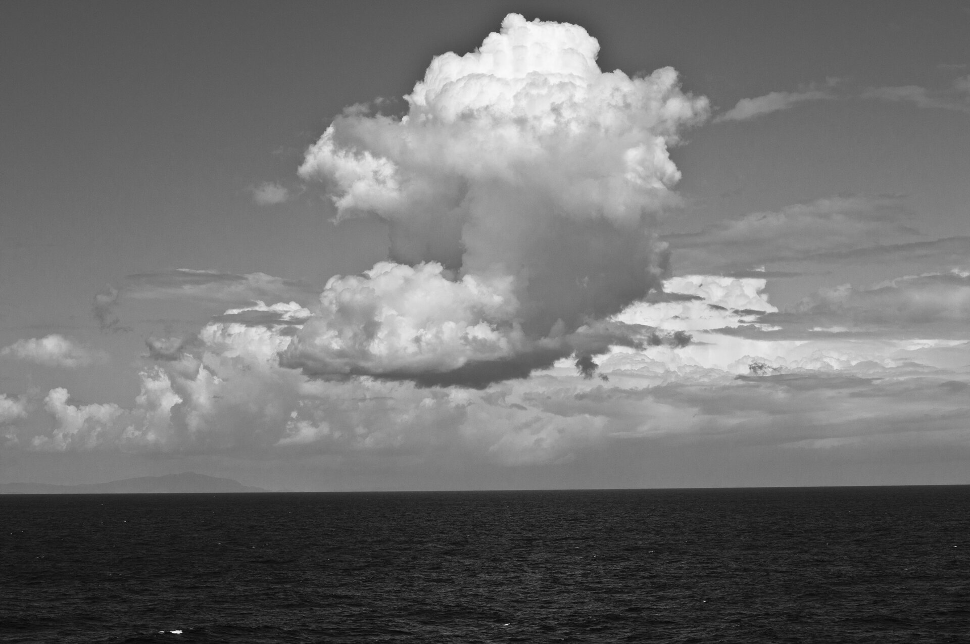 Ilustracja przedstawia fotografię „Niebo II”, autorstwa Anny Koli. Czarno‑białe zdjęcie ukazuje pejzaż morski. W centrum, na tle nieba unosi się duży obłok poszarpanej, białej chmury. Poniżej znajduje się wąski pas ciemnego, płaskiego morza. Nad horyzontem rysuje się szary kształt lądu. Praca skomponowana jest statycznie.