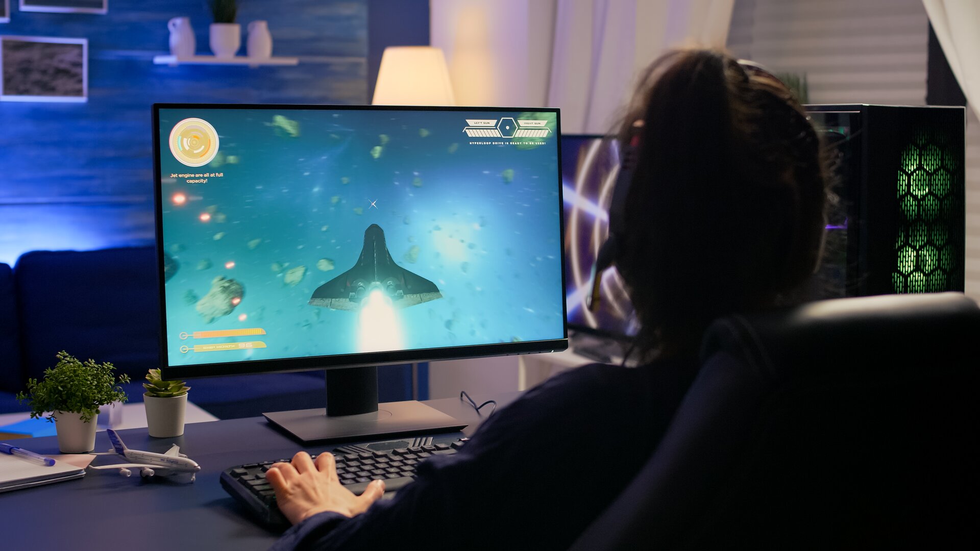 Zdjęcie przedstawia osobę siedzącą przed ekranem komputera. Ma założone słuchawki i trzyma dłonie na klawiaturze. Na ekranie widoczna jest gra polegająca na lataniu statkiem kosmicznym.