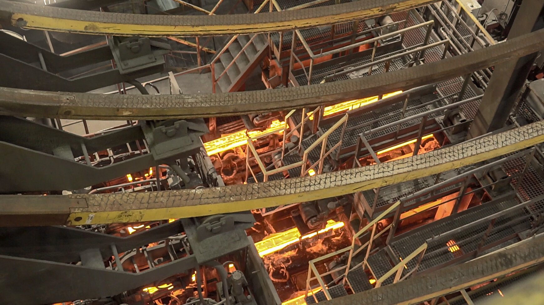 Na zdjęciu widać odlewane wlewki po wyjściu ze strefy wtórnego chłodzenia na urządzeniu COS (Ciągłe Odlewanie Stali). W centralnej części zdjęcia znajduje się dwa zakrzepnięte pasma rozgrzanej stali. Pasma mają kolor pomarańczowo złoty. Pasma znajdują się w indywidualnych korytach równoległych względem siebie. W tle zdjęcia widać wnętrze dużego obiektu przemysłowego.
