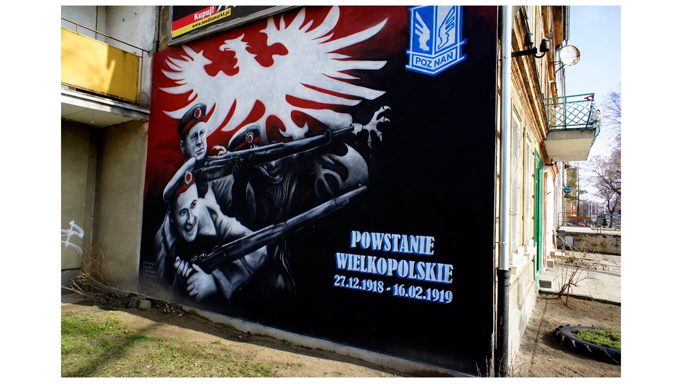 Fotografia barwna przedstawiająca mural upamiętniający powstanie wielkopolskie w Ostrowie Wielkopolskim na jednym z domów. W lewym górnym roku biały orzeł na czerwonym tle. Niżej dwóch żołnierzy z karabinami. Po prawej napis: Powstanie wielkopolskie, 27.12.1918–16.02.1919.