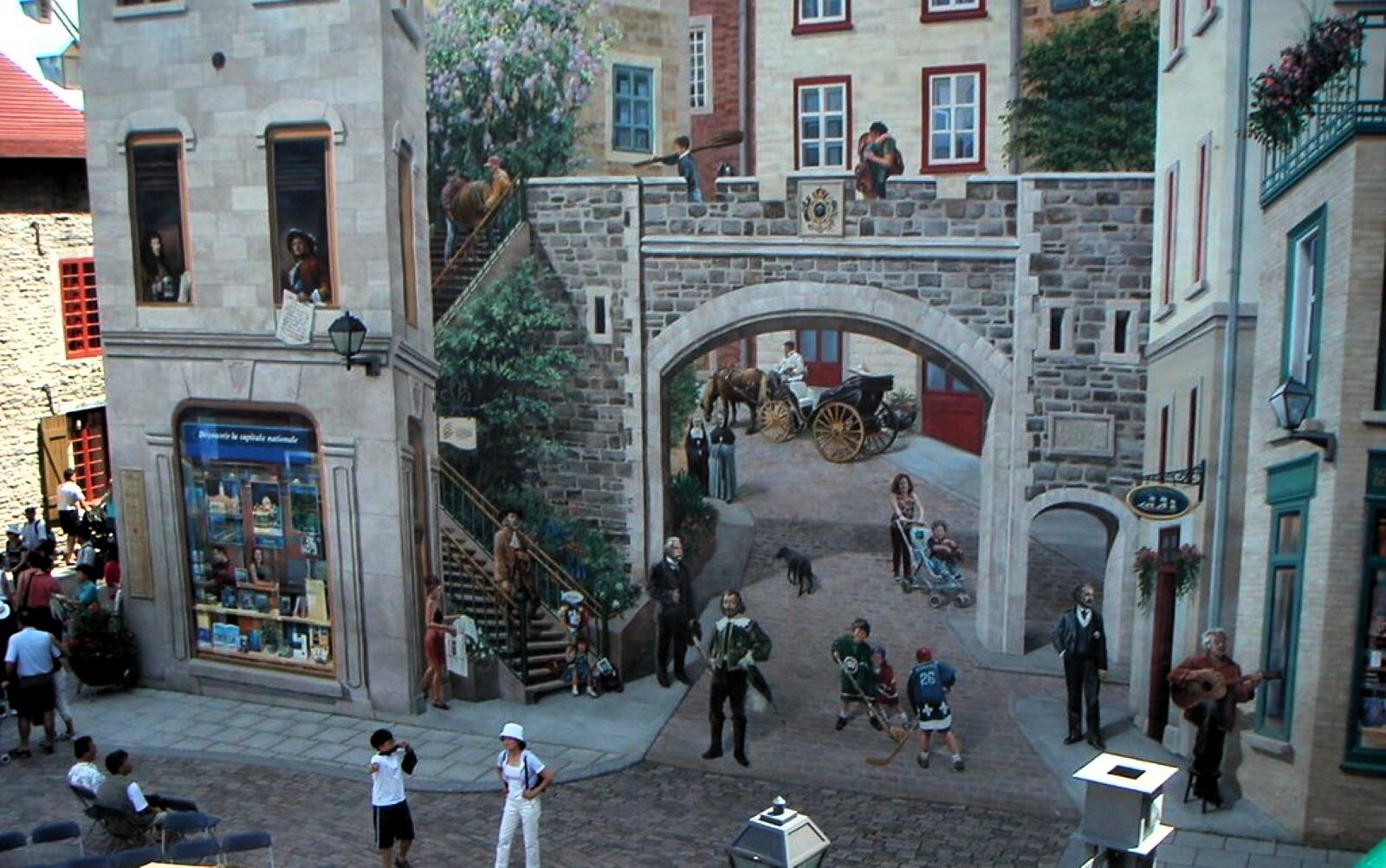 Zdjęcie przedstawia mural wykonany w mieście Quebec w Kanadzie. Jest iluzjonistyczny, bo namalowany na płaskim murze stwarza iluzję miejskiego zaułka z wejściem w małą, uroczą uliczkę. Są tam  ludzie z różnych epok; mężczyzna w ubraniu dworskim, uliczny śpiewak z gitarą, chłopcy w sportowych strojach grający w hokeja i całująca się para na kamiennym murze, a także inne osoby.