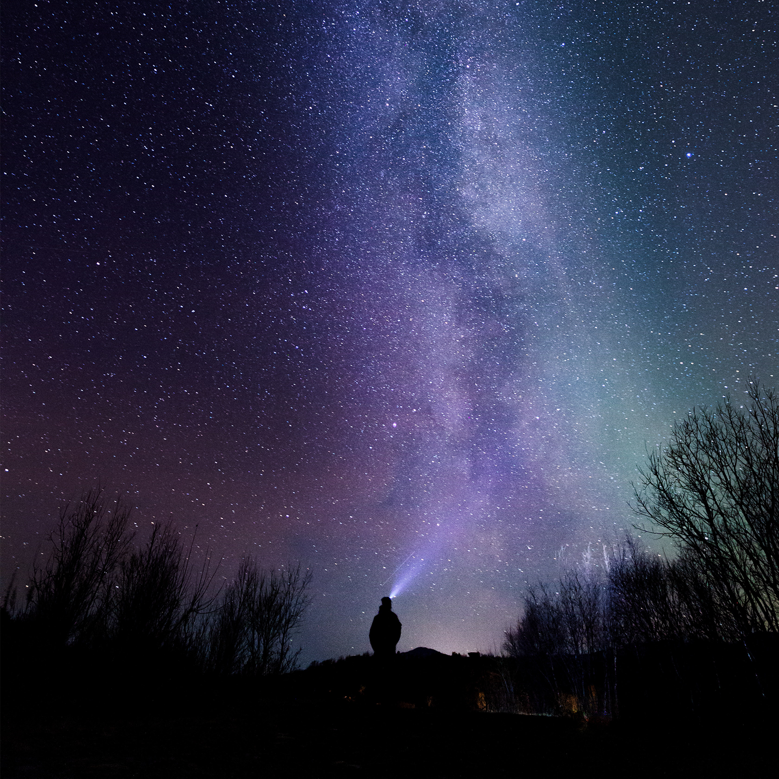 Zdjęcie przedstawia rozgwieżdżone niebo z widoczną mleczną drogą. Widoczny jest też człowiek, znad głowy którego wydobywa się snop światła skierowany w niebo. 