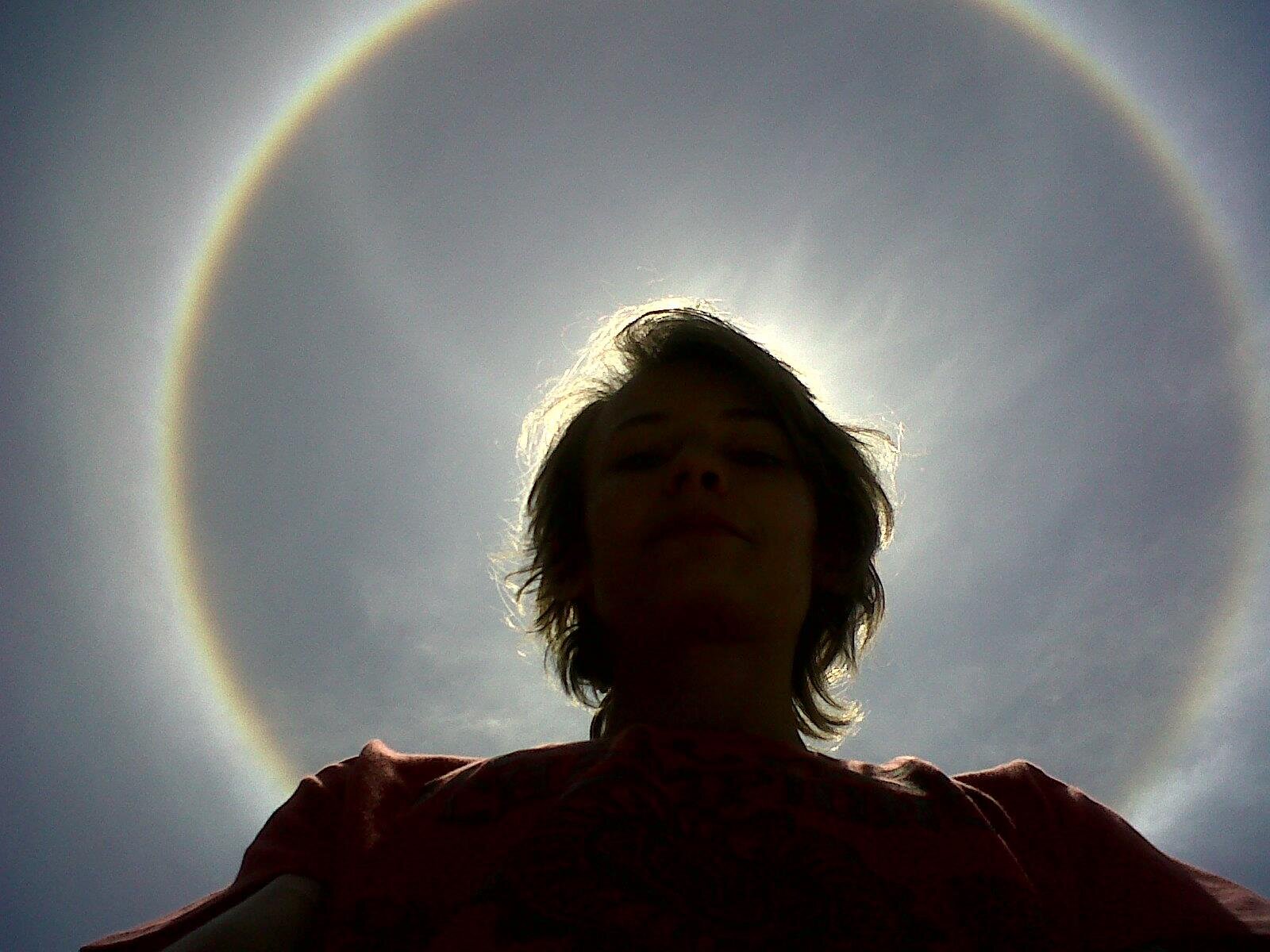 Fotografia barwna przedstawiająca zjawisko atmosferyczne halo. Na tle pierzastych chmur głowa młodego mężczyzny otoczona świetlistym, okręgiem wyglądającym jak tęcza.