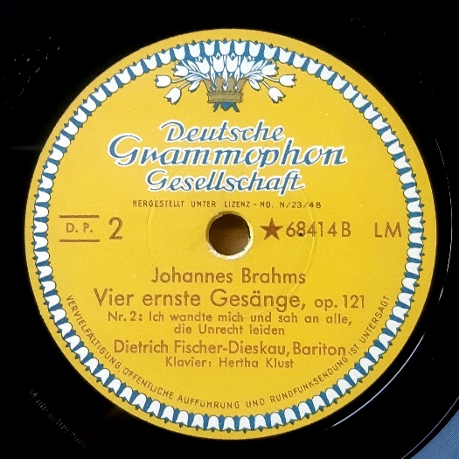 Fotografia przedstawia zbliżenie na fragment płyty winylowej z żółtą etykietą i logo Deutsche Grammophon [czytaj: dojcze gramofon]. Poniżej znajduje się napis: Johannes Brahams, Vier ernste Gesänge [czytaj: johanes brahams, wie ernste gesenje] , op.121, Nr. 2: Ich wandte mich und sah an alle  [czytaj: iś wande miś un sah nach ale], die Unrecht leiden  [czytaj: di unreśt lajden], Dietrich Fischer- Dieskau  [czytaj: ditriś fisszar diksał], Bariton, Klavier: Herth Klust  [czytaj: klawier: hert klus]. Logo składa się z bukietu tulipanów. Umieszczone zostało w górnej części etykiety.