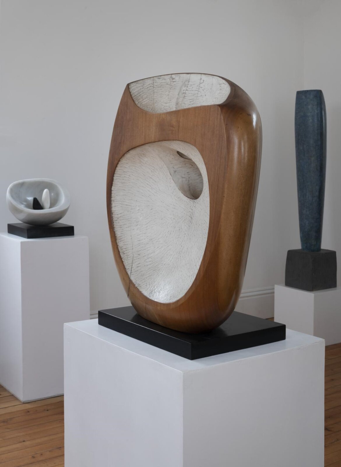 Na fotografii znajduje się rzeźba, ustawiona na białym cokole. Rzeźba to drewniany, obły element w brązowym kolorze, a środkowa część jest biała i ma inną strukturę. Z tyłu znajdują się jeszcze dwie rzeźby, również na cokole.