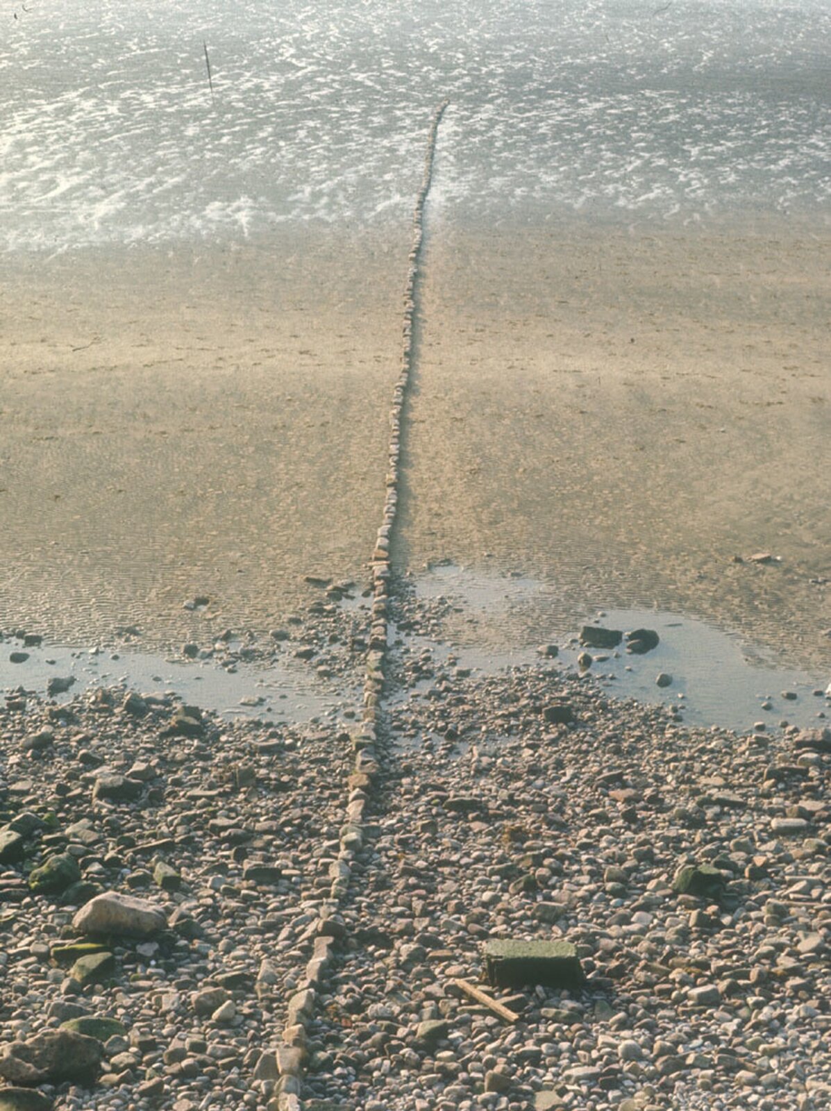 Ilustracja przedstawia przykład dzieła Andy'ego Goldworthy'ego. Na fotografii widzimy fragment plaży. Plaża jest podzielona na 3 części: część kamienistej plaży, część piaszczystej plaży oraz woda. Poprzez środek fotografii przebiega linia. Widoczny jest wyraźny odstęp pomiędzy poszczególnymi częściami plaży. Przez całą długość fotografii jest ułożona z kamieni cienka linia przebiegająca od krawędzi zdjęcia na środku, od dołu do początku brzegu morza.