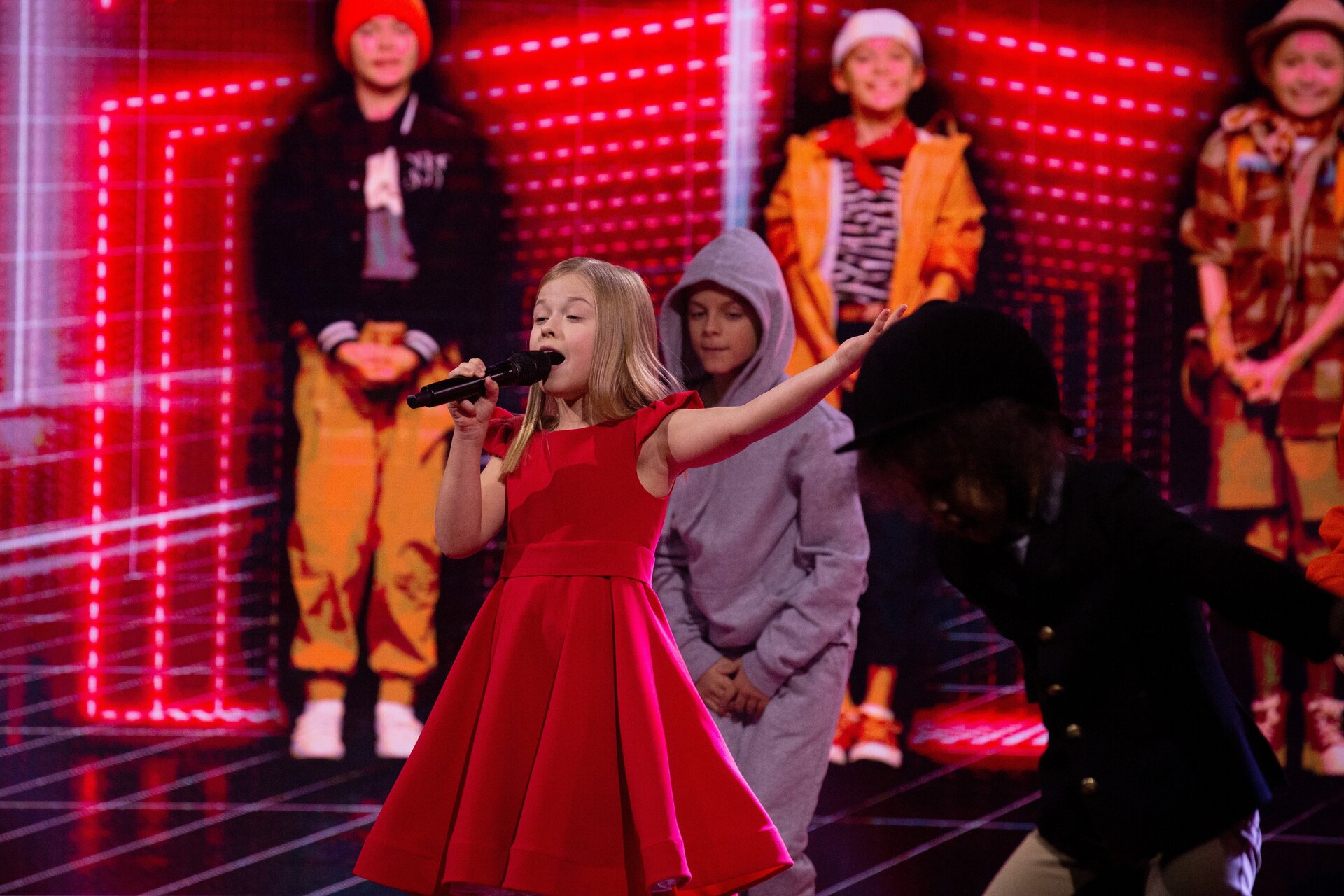 Fotografia przedstawia młodą dziewczynkę o długich blond włosach, ubraną w długą czerwoną sukienkę. Dziewczynka w jednej dłoni trzyma mikrofon, a drugą ma wyciągniętą ku górze. Za piosenkarką stoi grupa choreograficzna tańcząca w rytm muzyki.