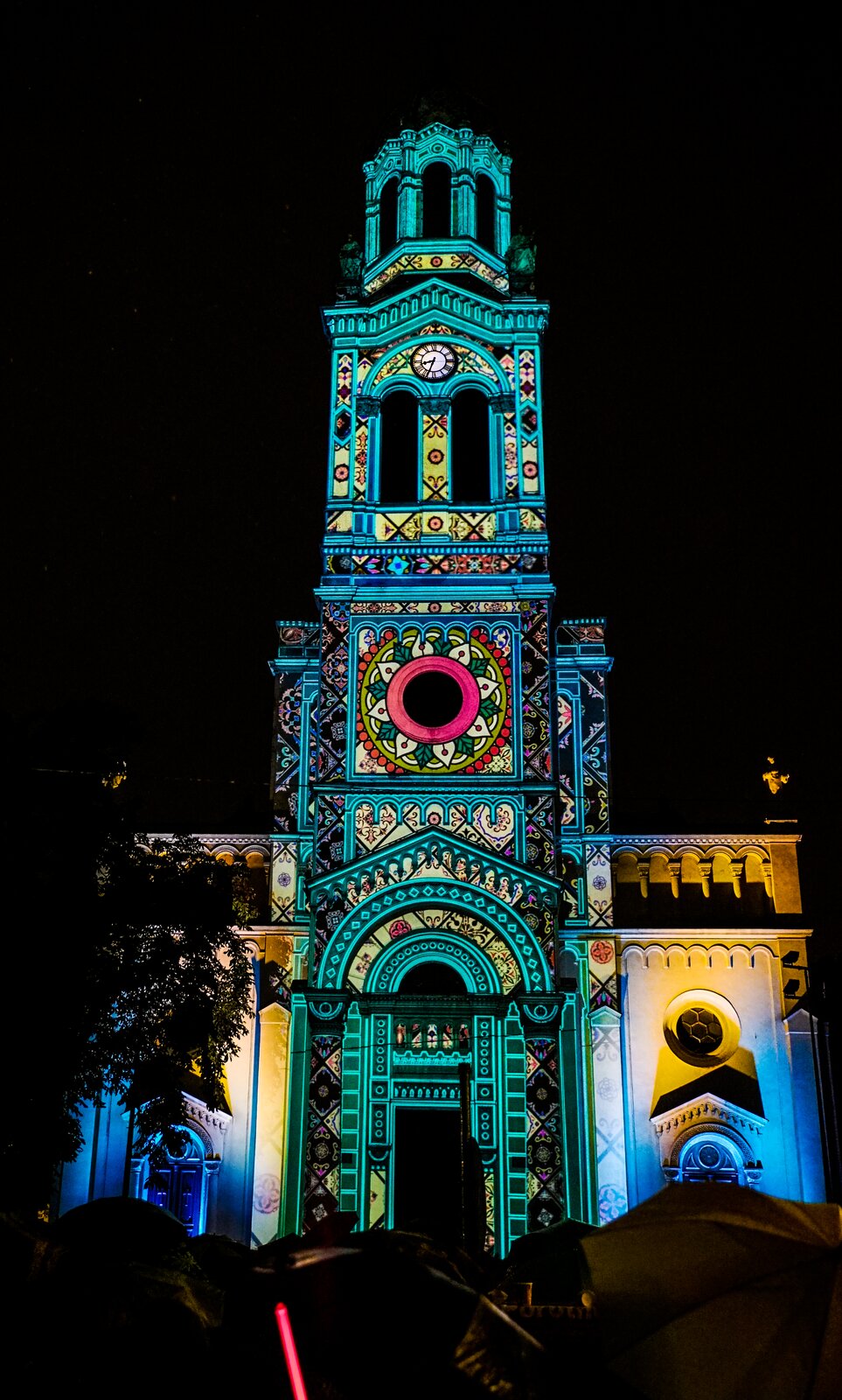 Na zdjęciu znajduje się podświetlony budynek z wieżą. Na jego fasadzie wyświetlone zostały  kolorowe, geometryczne wzory. Przed budynkiem stoi wysokie drzewo i ludzie z parasolami. Tło stanowi ciemne nocne niebo. Fotografia jest ciemna, jedynie budynek jest rozświetlony.