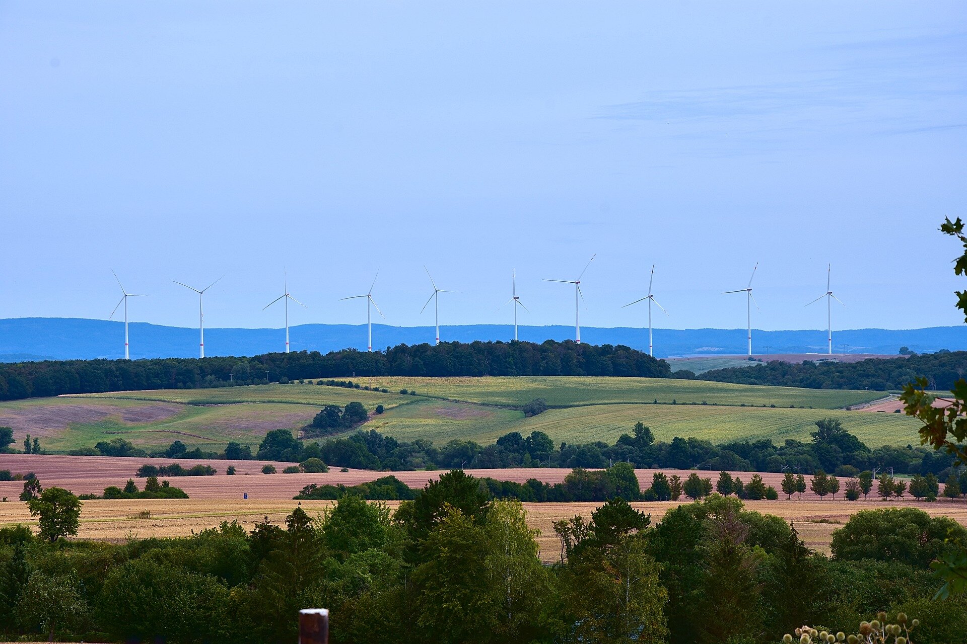 Zdjęcie przedstawia elektrownię wiatrowa znajdującą się na wzniesieniu. Dookoła rosną drzewa, są łąki. 