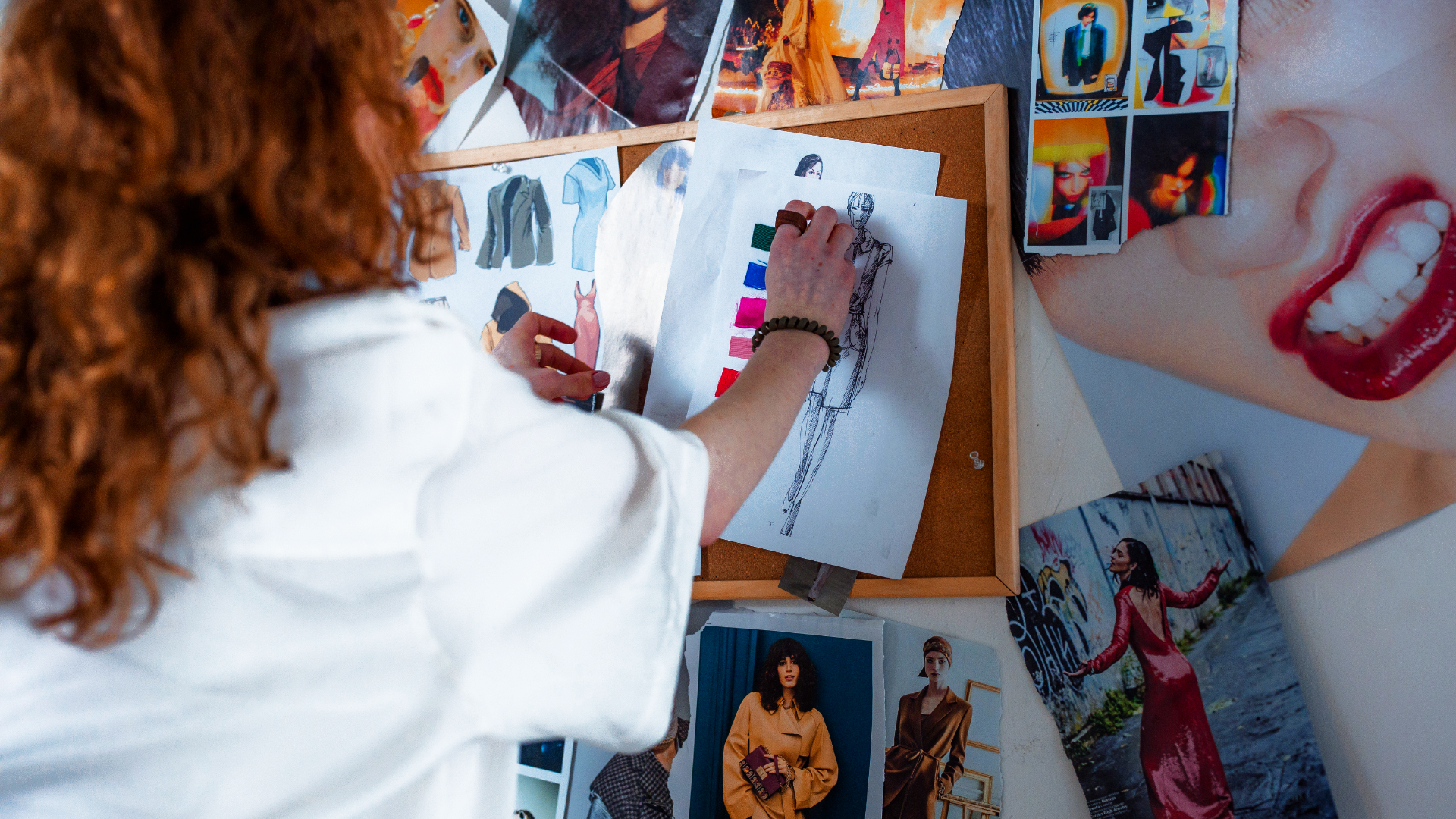 Zdjęcie przedstawia kobietę/kostiumografkę (jej twarz jest niewidoczna, kobieta stoi tyłem do kadru), która na ścianie pełnej wycinków z kolorowych gazet przypina rysunek sylwetki z paletą kolorów.