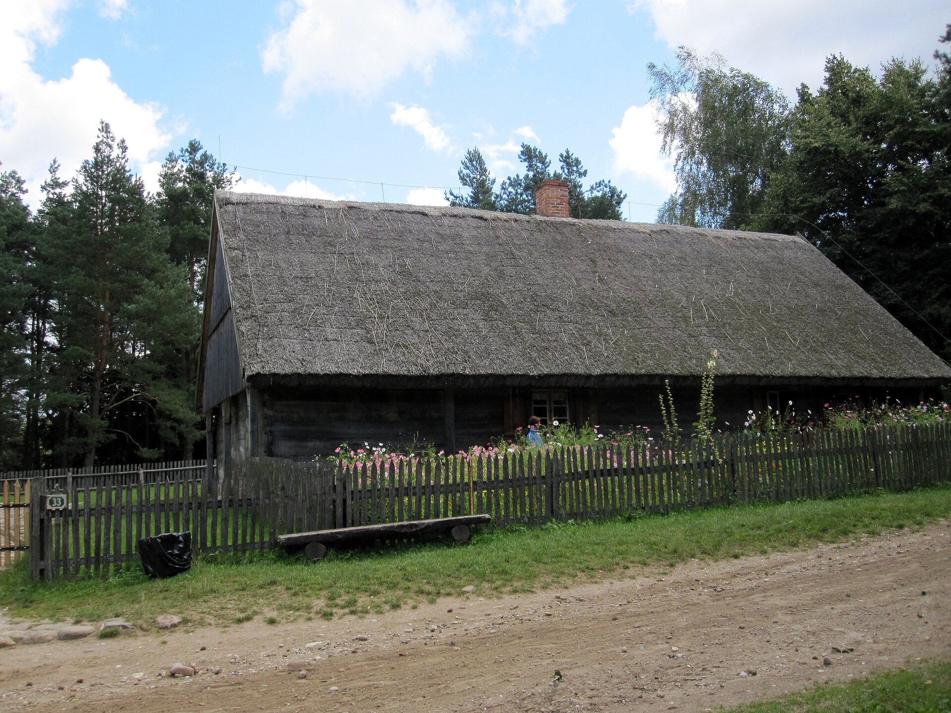Zdjęcie przedstawia odrestaurowaną tradycyjną drewnianą chałupę na terenie skansenu. Na pierwszym planie droga gruntowa, dalej płot, przy którym rosną kwiaty. Za domem linia drzew.