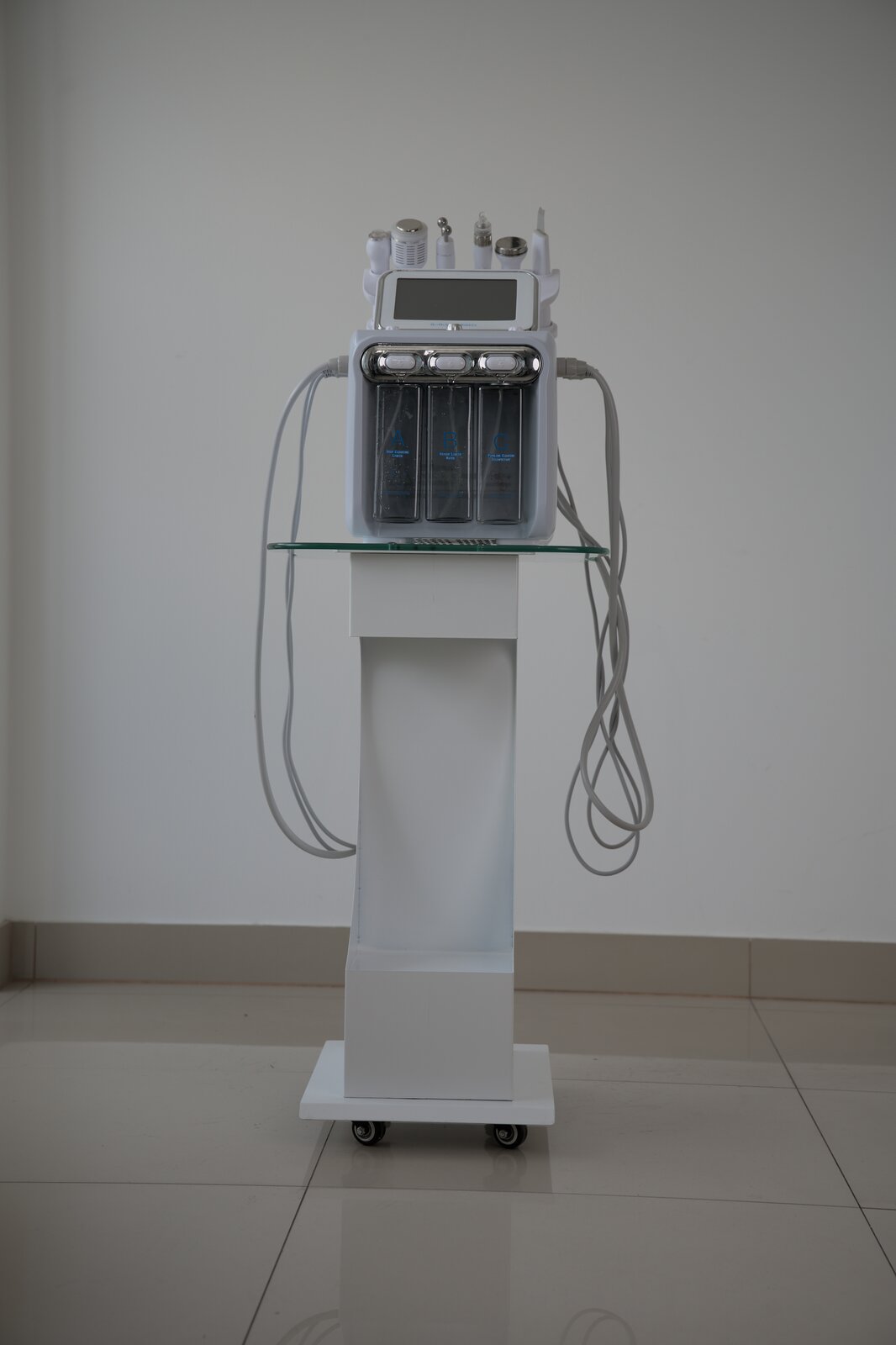Zdjęcie przedstawia urządzenie wielofunkcyjne do zabiegów kosmetycznych z zastosowaniem fali radiowej, ultradźwięków i peelingu kawitacyjnego. Urządzenie jest na wysokim stoliku na kółkach. Składa się z korpusu, na którym jest ekran dotykowy. Do korpusu przyłączone są przewodami głowice o różnych kształtach do zabiegów kosmetycznych.