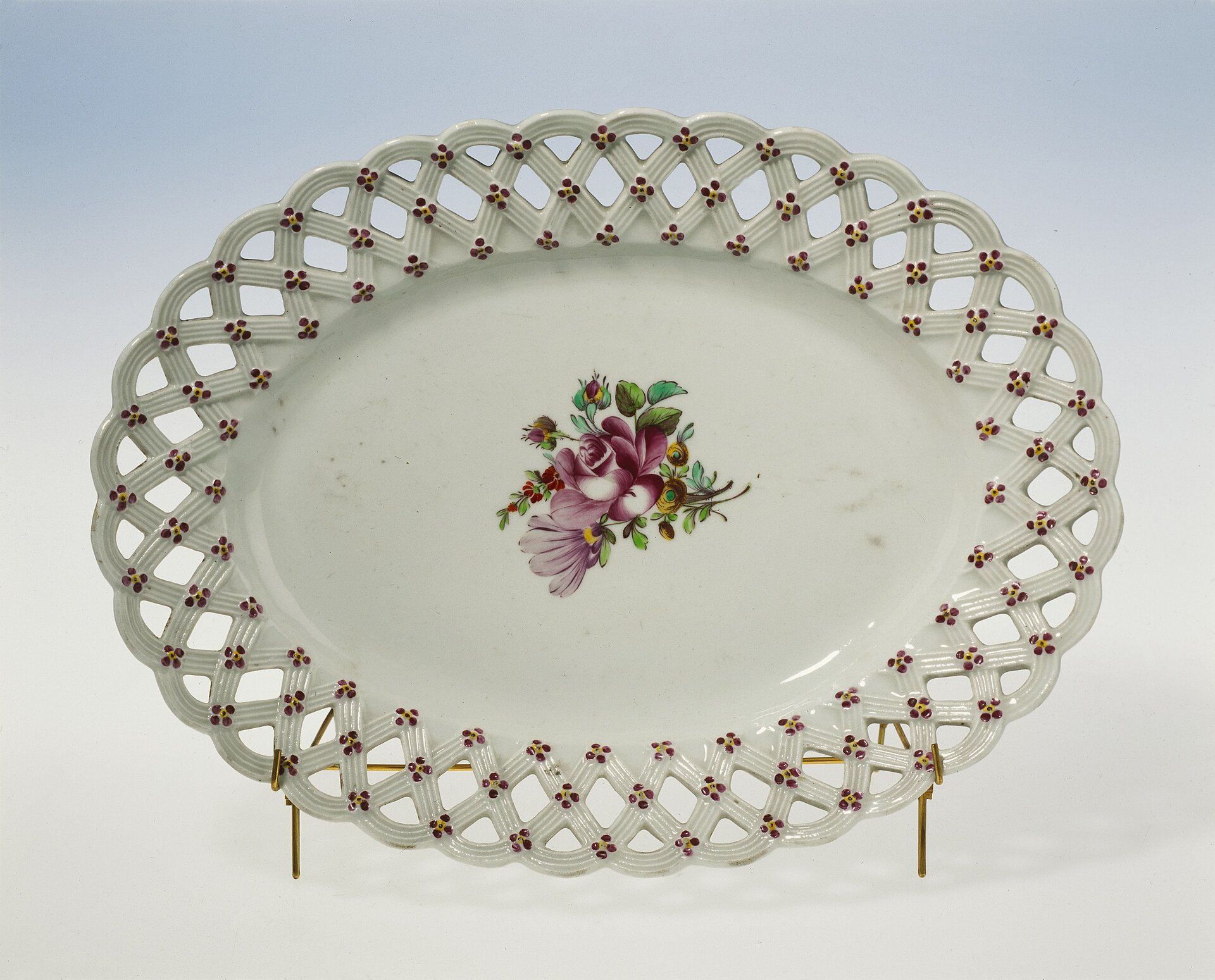 Kliknij, aby powiększyć Zdjęcie przedstawia wytwór manufaktury koreckiej w postaci owalnego talerza w ornamentyce plecionkowej z motywem kwiatowym. Na środku naczynia jest charakterystyczny dla zakładu wzór kwiatowy róży w rozkwitnieniu.