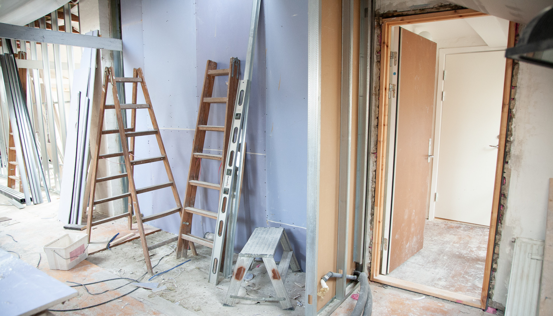 Zdjęcie przedstawia wnętrze domu w czasie remontu. Remont jest w początkowym etapie. Widoczna jest obsadzona futryna drzwi. Po lewej znajduje się konstrukcja z aluminiowych profili. W pomieszczeniu znajdują się dwie drabiny.
