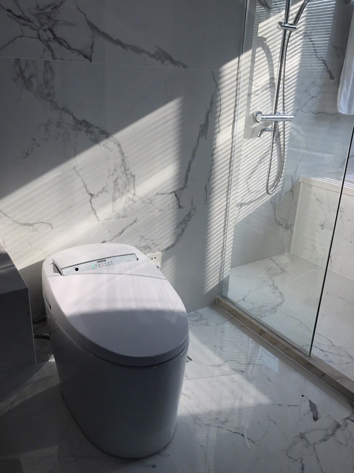 Zdjęcie przedstawia łazienkę z elektryczną muszlą klozetową w kolorze białym, z panelem sterującym umieszczonym w tylnej części deski. Na panelu widoczny jest czarny przycisk oraz skala. Na prawo od muszli klozetowej widoczna jest przeszklona kabina prysznicowa. Ściany i podłoga pokryte są dużymi płytkami marmurowymi.