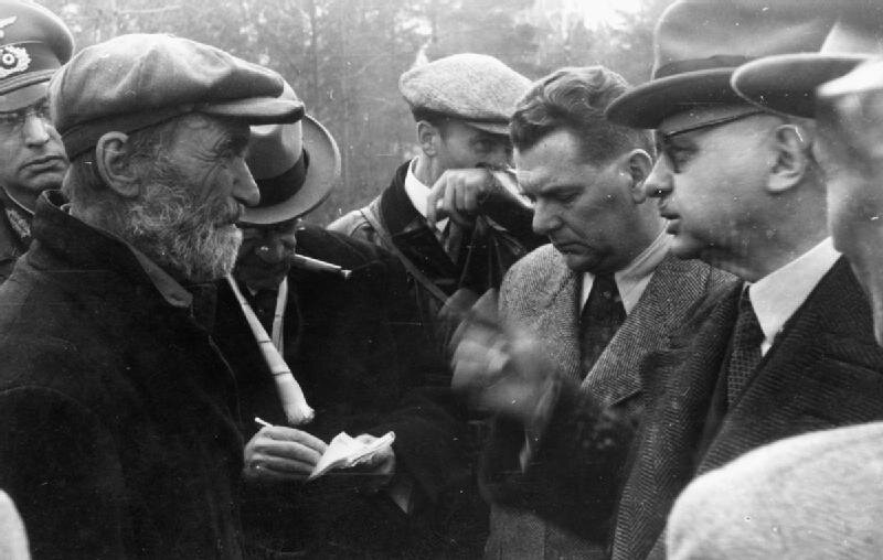 Ilustracja przedstawia kilku mężczyzn stojących w okręgu i rozmawiających. Jeden z mężczyzn to oficer niemiecki, reszta ubrana jest po cywilnemu. Na pierwszym planie starszy mężczyzną z siwą brodą, pomarszczoną twarzą rozmawia z pucułowatym dygnitarze, w okularach i okrągłych okularach.