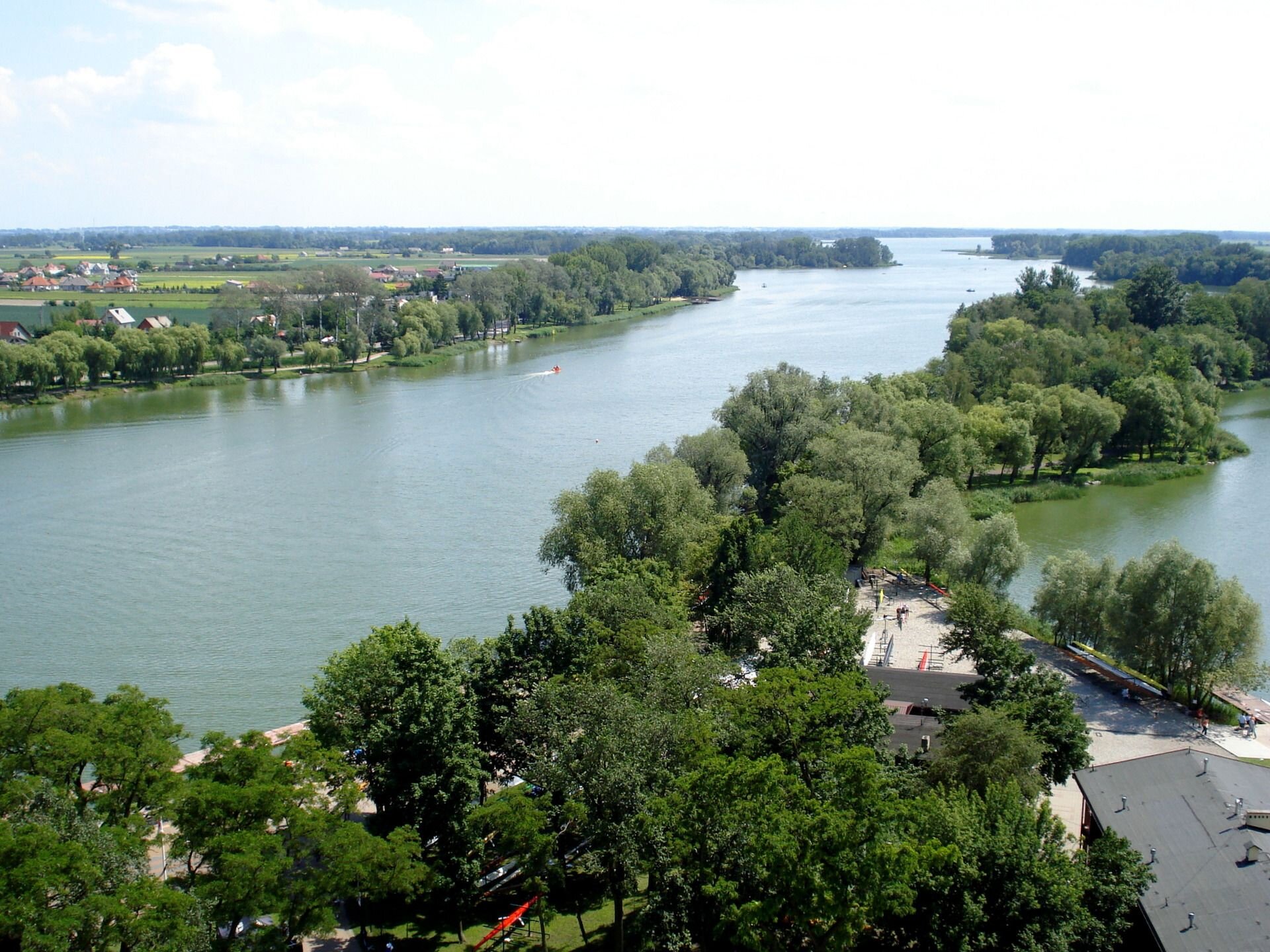 Zdjęcie przedstawia jezioro Gopło, które jest rynnową formą polodowcową. Rynnowaty podłużny kształt jeziora jest ukazany na zdjęciu. Brzegi jeziora porośnięte są drzewami ,wśród których są dachy domów.