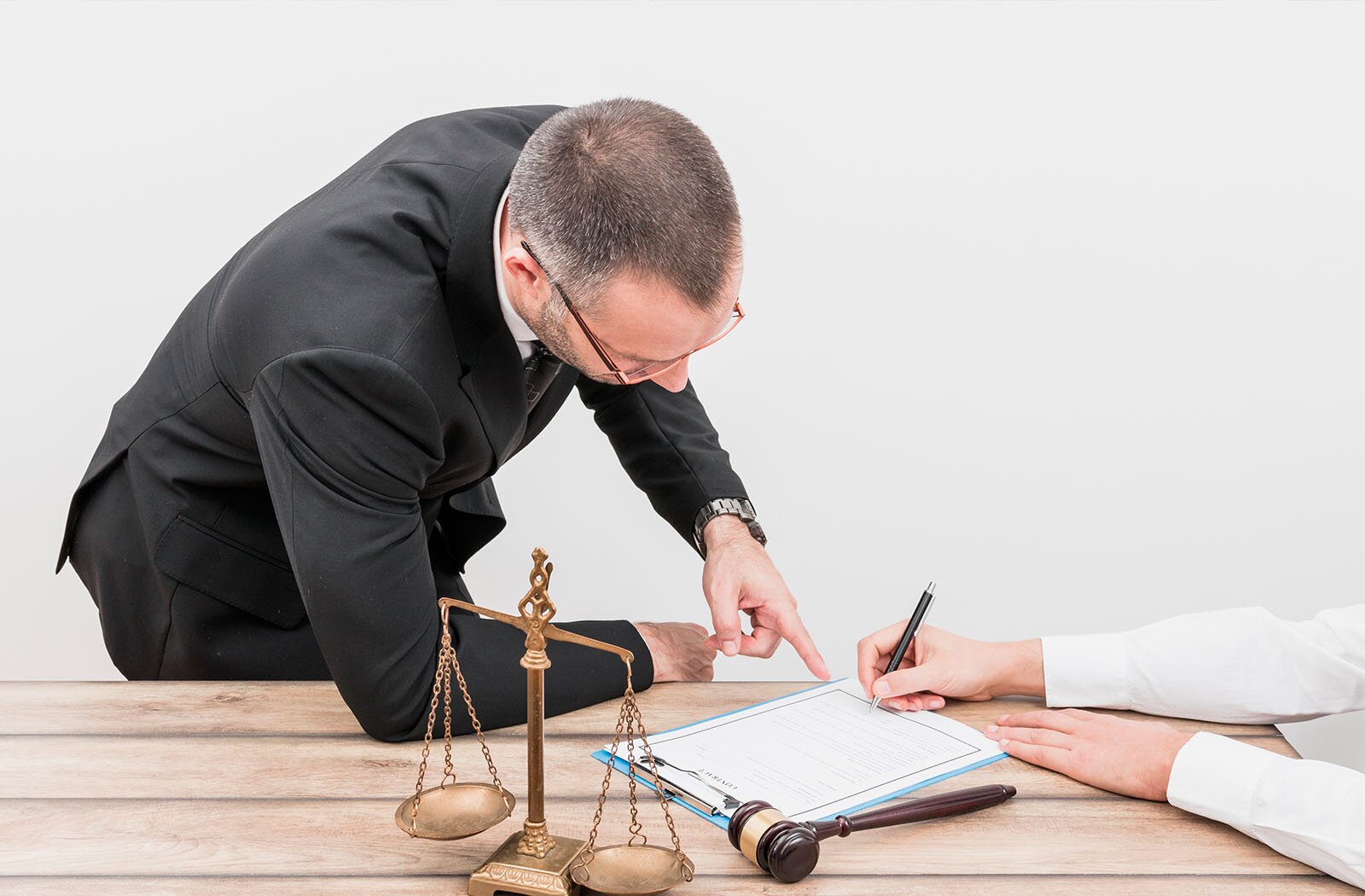 Na zdjęciu widoczny jest pochylający się mężczyzna w garniturze, oparty prawym łokciem o blat biurka. Palcem wskazującym lewej ręki dotyka rogu kartki leżącej na blacie. Po prawej stronie widoczne są dwie ręce. Prawa dłoń trzyma długopis uniesiony nad dolną krawędzią strony, tuż obok palca mężczyzny, a lewa leży na biurku. Na blacie widoczny jest również młotek sędziowski oraz złota waga szalkowa.