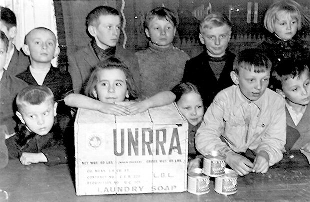 Fotografia przedstawia grupę dzieci stojących przy ladzie gdzie wydawane są paczki z żywnością. Na jednej z paczek napis: UNRRA.