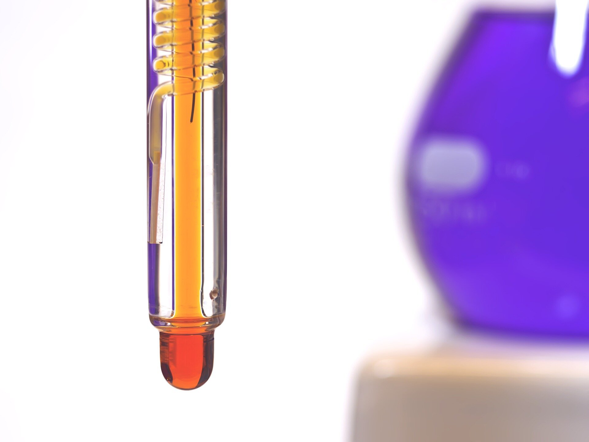 Zdjęcie przedstawiające szklane, podłużne naczynie, we wnętrzu naczynia znajduje się szklana spirala, wypełniona pomarańczową cieczą. W tle fragment kolby wypełnione fioletowym roztworem.