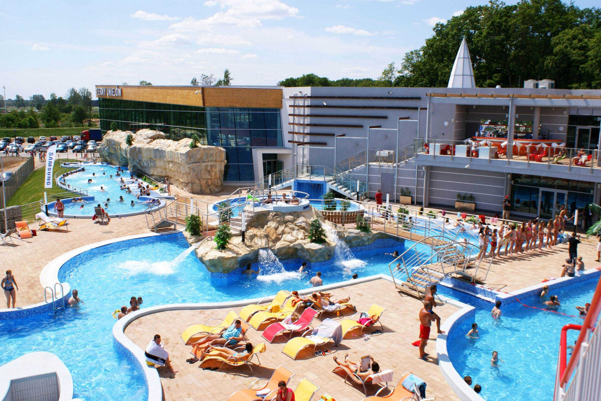 Zdjęcie przedstawia przed rozległym budynkiem ośrodka z częściowo przeszkloną ścianą znajdują się liczne, niekiedy połączone baseny, w których kąpią się ludzie. Pośrodku dodatkowo jacuzzi, fontanny, mostki.