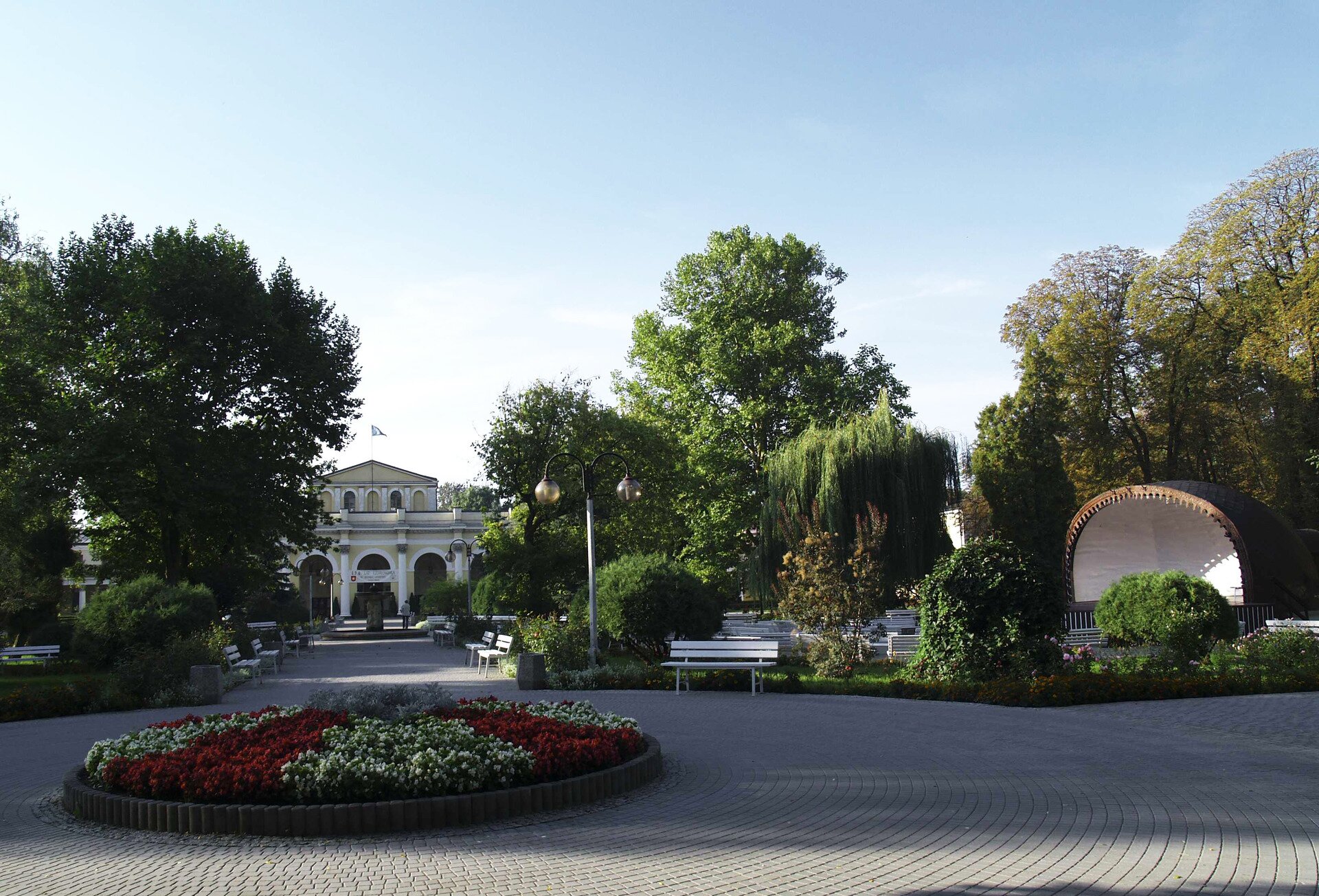 Fotografia prezentująca fragment parku uzdrowiskowego w Busku Zdroju. Na pierwszym planie widoczny okrągły klomb, od którego prowadzi aleja w kierunku budynku z kolumnami.  Po prawej stronie mini amfiteatr otoczony drzewami przed którym znajdują się ławeczki.
