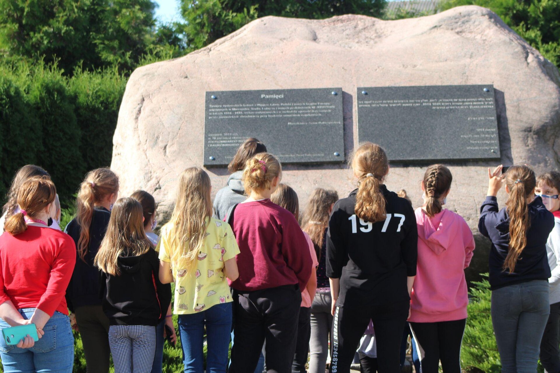 Zdjęcie przedstawia duży, jasny kamień, do którego zostały przytwierdzone dwie, ciemne tablice pamiątkowe. Napisy na tablicach są jasne i nieczytelne. Przed kamieniem, tyłem do obiektywu, stoją młode osoby - czternaście uczniów, którzy ubrani są w kolorowe ubrania. Jedna z osób robi zdjęcie tablicy. W tle widoczne są zielone krzewy.