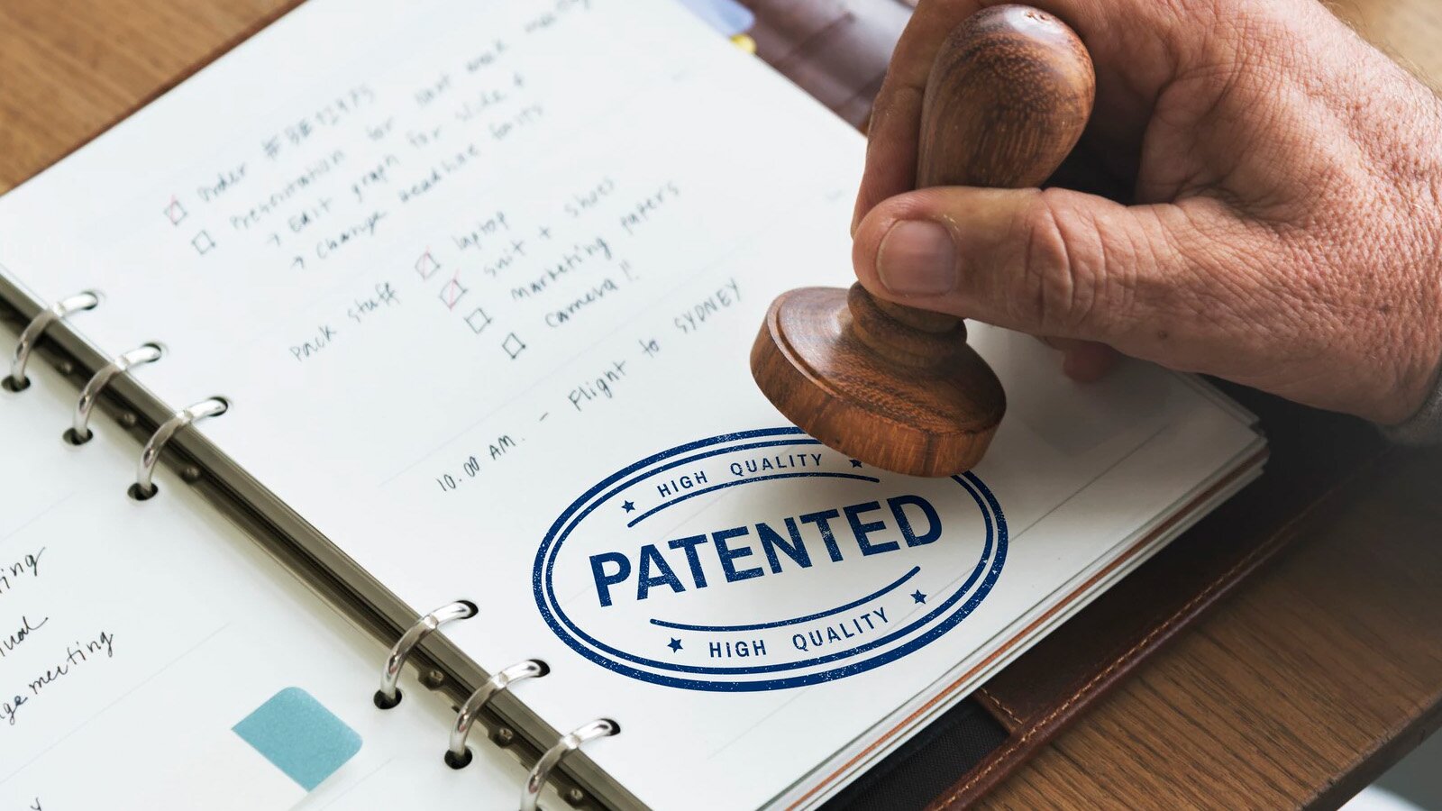 Zdjęcie przedstawia otwarty notes, tak zwany kołonotatnik, wypełniony notatkami. Nad notesem znajduje się uniesiona męska dłoń trzymająca drewniany stempel. Przed chwilą tym stemplem został odciśnięty widoczny u dołu kartki wzór z napisem PATENTED.