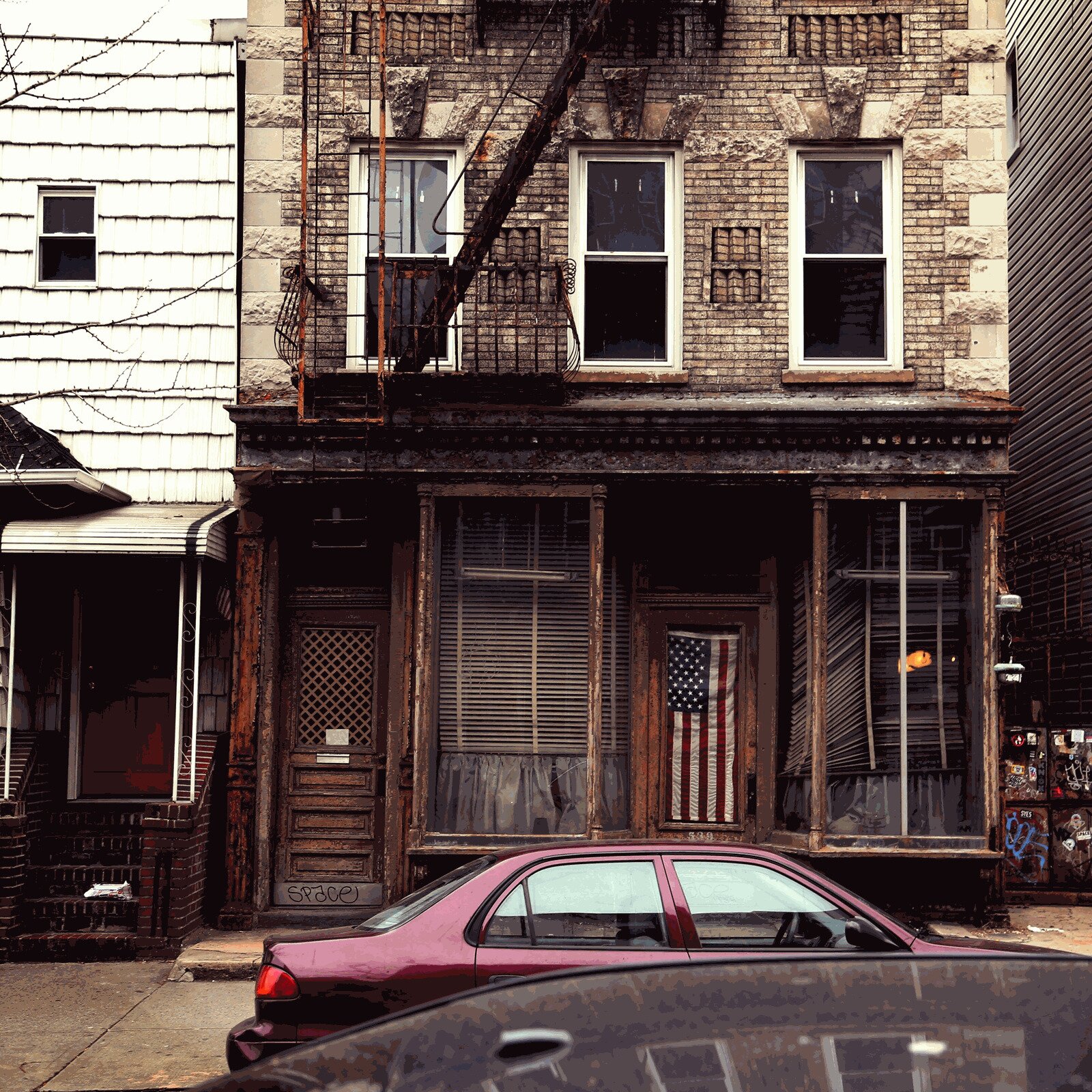 Zdjęcie przedstawia piętrowy, ceglany budynek, z drewnianym, zrujnowanym gankiem. W jego oknie zawieszona jest flaga Stanów Zjednoczonych. Przed domem stoją zaparkowane samochody.