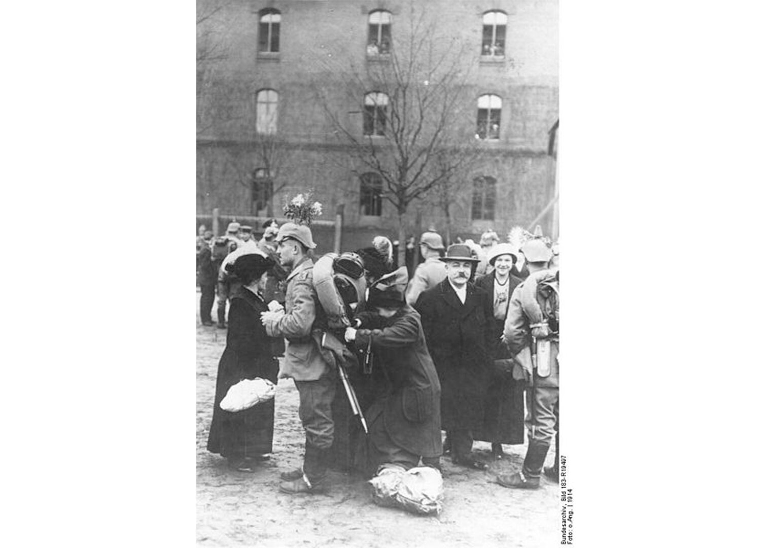 Zdjęcie czarno-białe przedstawiające niemieckich żołnierzy i żegnających ich krewnych. Żołnierze w pełnym umundurowaniu rozmawiają z najbliższymi, kobiety trzymają zapakowane tobołki, pakują coś do żołnierskiego plecaka. Rodziny uśmiechają się.. 