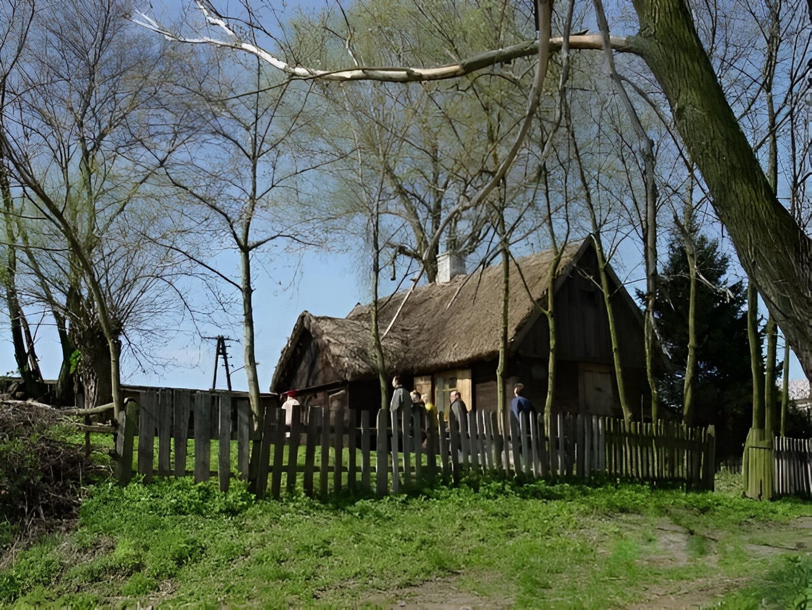 Fotografia przedstawia drewnianą chatę ze słomianym dachem ogrodzoną drewnianym płotem. Wokół chaty drzewa i ludzie.