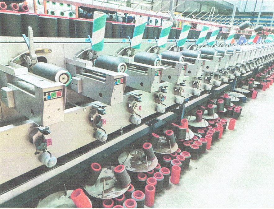 Zdjęcie przedstawia przewijarkę. Maszyna składa się z licznych, takich samych stanowisk przewijania. Na każdym stanowisku zamontowana jest szpulka szarych nici. Przy zamontowanych szpulkach są panele sterujące. Na maszynie i pod maszyną stoją inne szpulki.