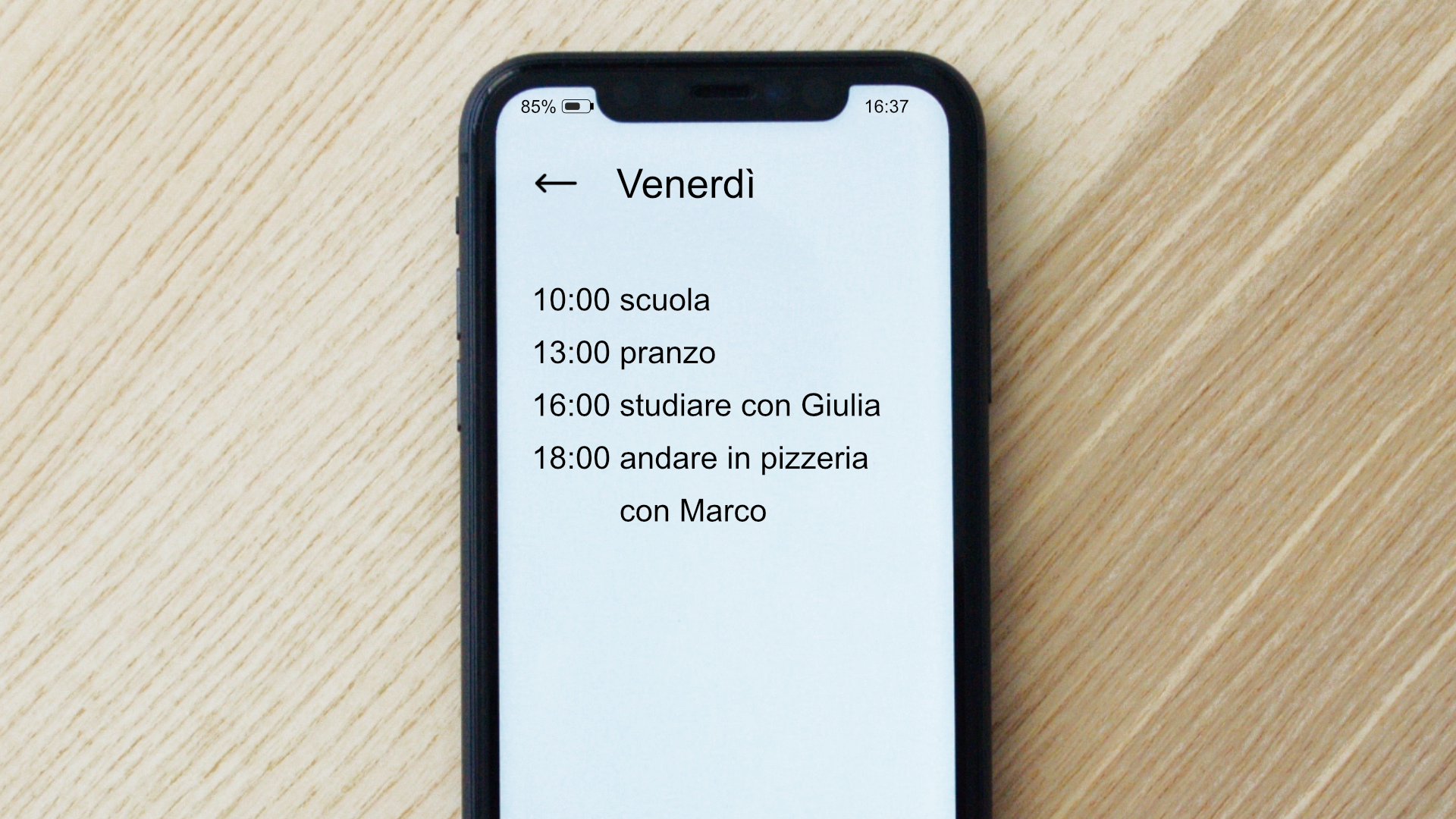 Zdjęcie przedstawia telefon komórkowy. Na ekranie telefonu znajduje się kalendarz z zapisanym planem dnia: Venerdì10:00 scuola13:00 pranzo16:00 studiare con Giulia18:00 andare in pizzeria con Marco