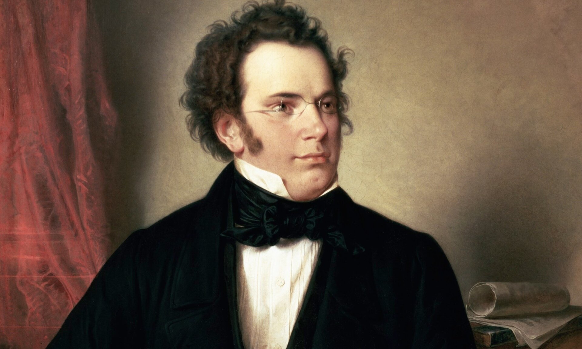 Zdjęcie przedstawia portret Franciszka Schuberta. Kompozytor ma okulary. Jego włosy są czarne i krótkie. Za nim znajduje się czerwona kotara.
