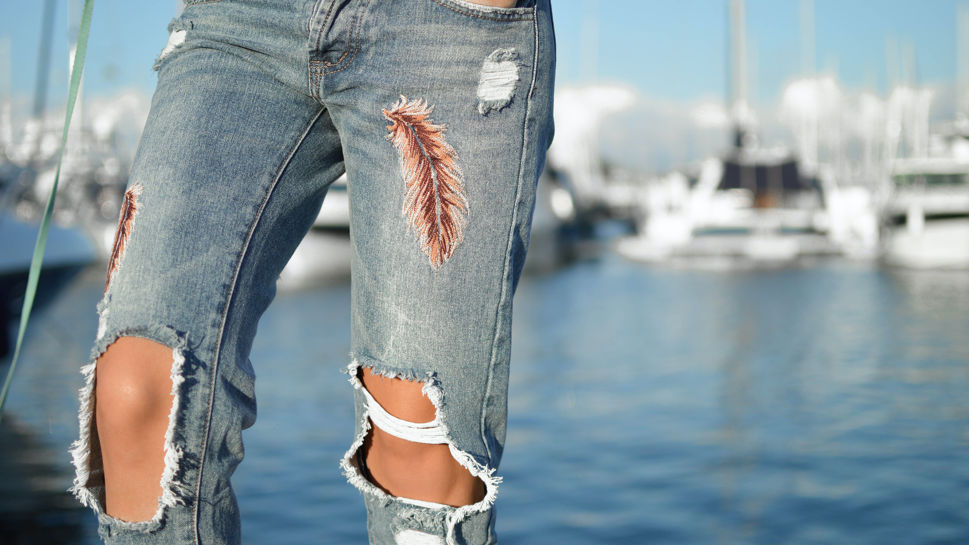 Zdjęcie przedstawia zbliżenie na nogi osoby która nosi dżinsy z dziurami na kolanach. Na spodniach, na wysokości ud, znajdują się również ilustracje ptasich piór.
