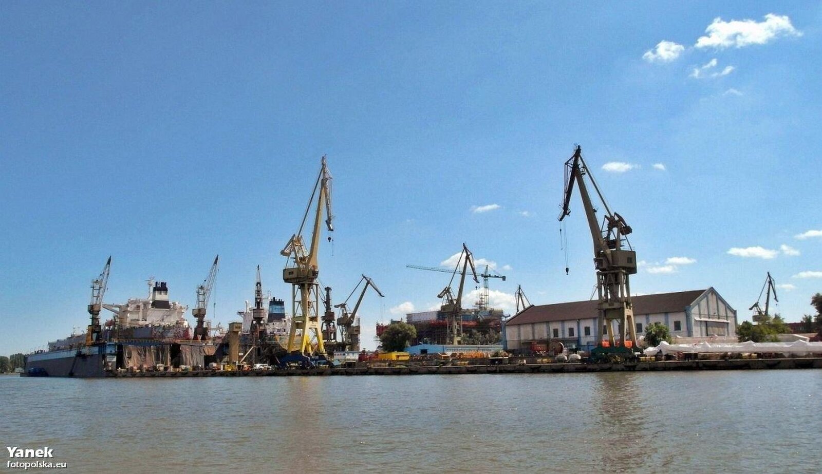 Zdjęcie przedstawiające Stocznię Remontową. Liczne wysokie dźwigi stojące tuż przy linii brzegowej, niskie zabudowania, magazyny.