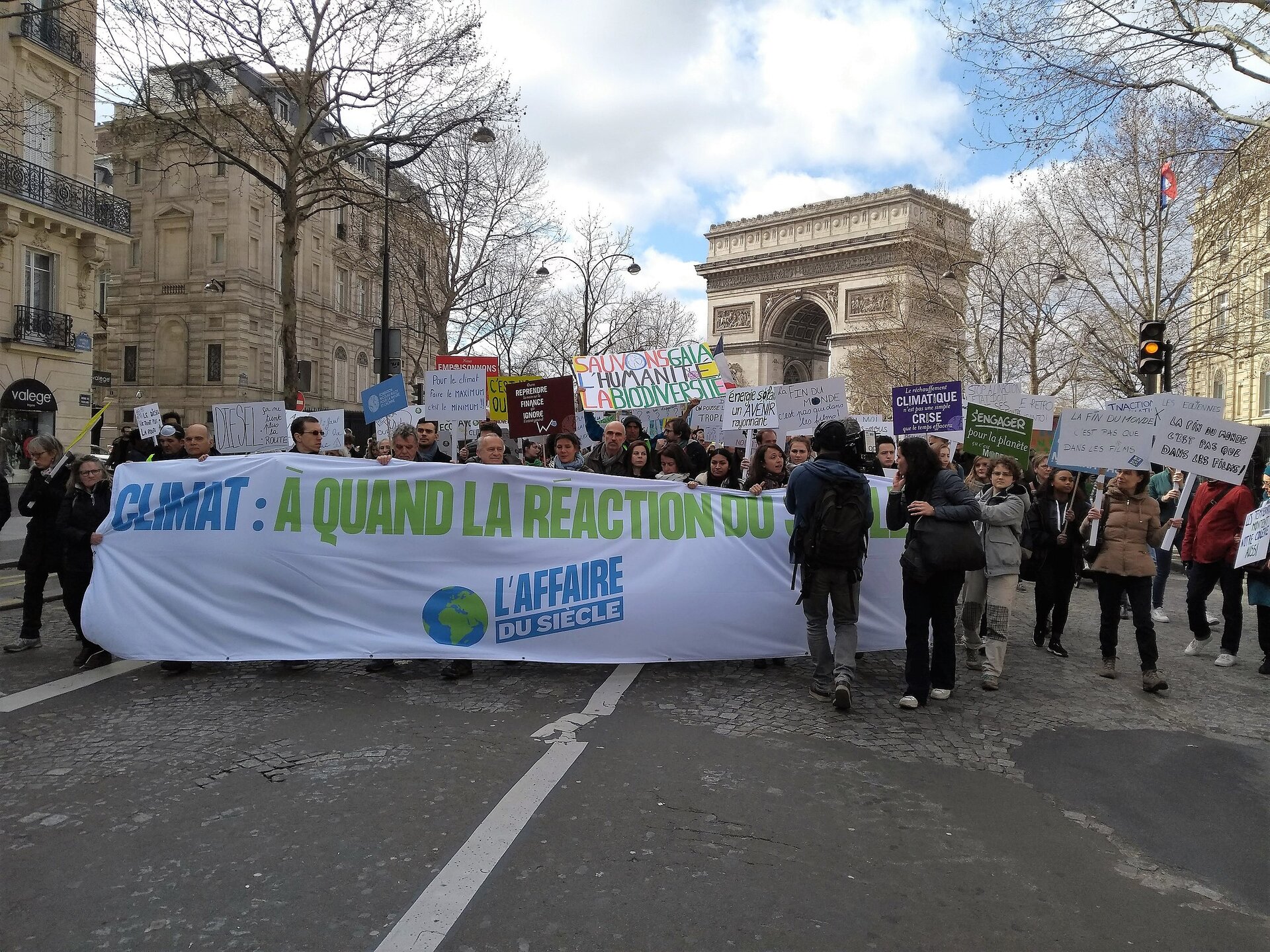 Ilustracja przedstawia grupę idących ulicą demonstrantów. Pierwszy rząd osób niesie długi transparent z napisem: Climat: à quand la réaction du.... 