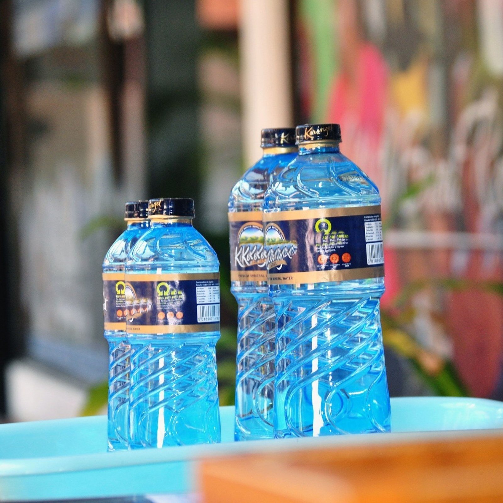 Zdjęcie przedstawia plastikowe butelki z wodą stojące w niebieskim  pojemniku przy lustrze. Butelka po lewej stronie jest mała, a ta po prawej stronie jest duża. W lustrze widoczne jest odbicie obu butelek.