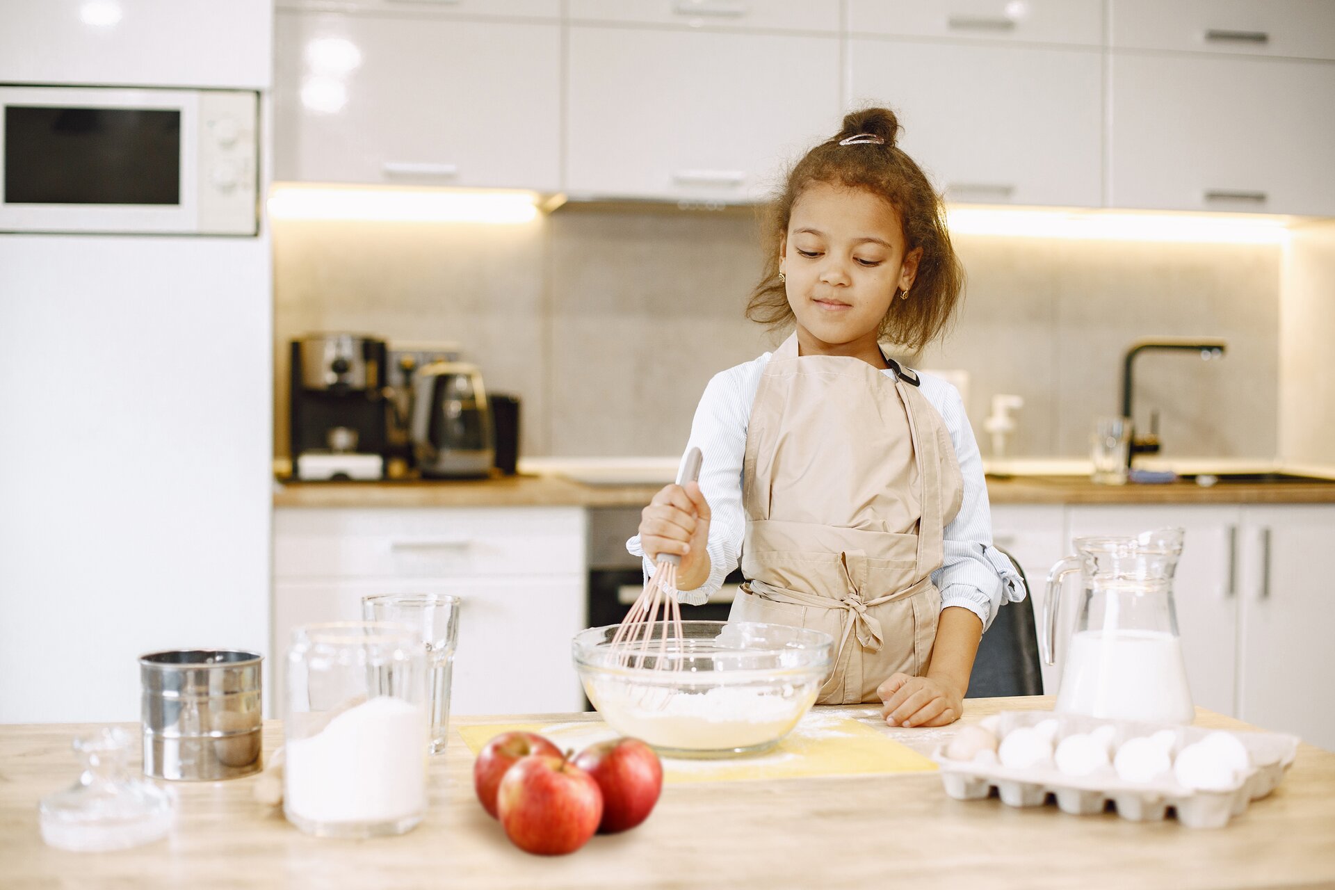 Zdjęcie przedstawia dziewczynkę w wieku wczesnoszkolnym, ubraną w fartuch. Dziecko stoi przy stole, w prawej ręce trzymając trzepaczkę, która jest zanurzona w misce z białym płynem. Na blacie stoi również dzbanek z mlekiem, jajka, jabłka, pojemnik z białym proszkiem oraz srebrna puszka. Za plecami dziewczynki widać zabudowę kuchenną.