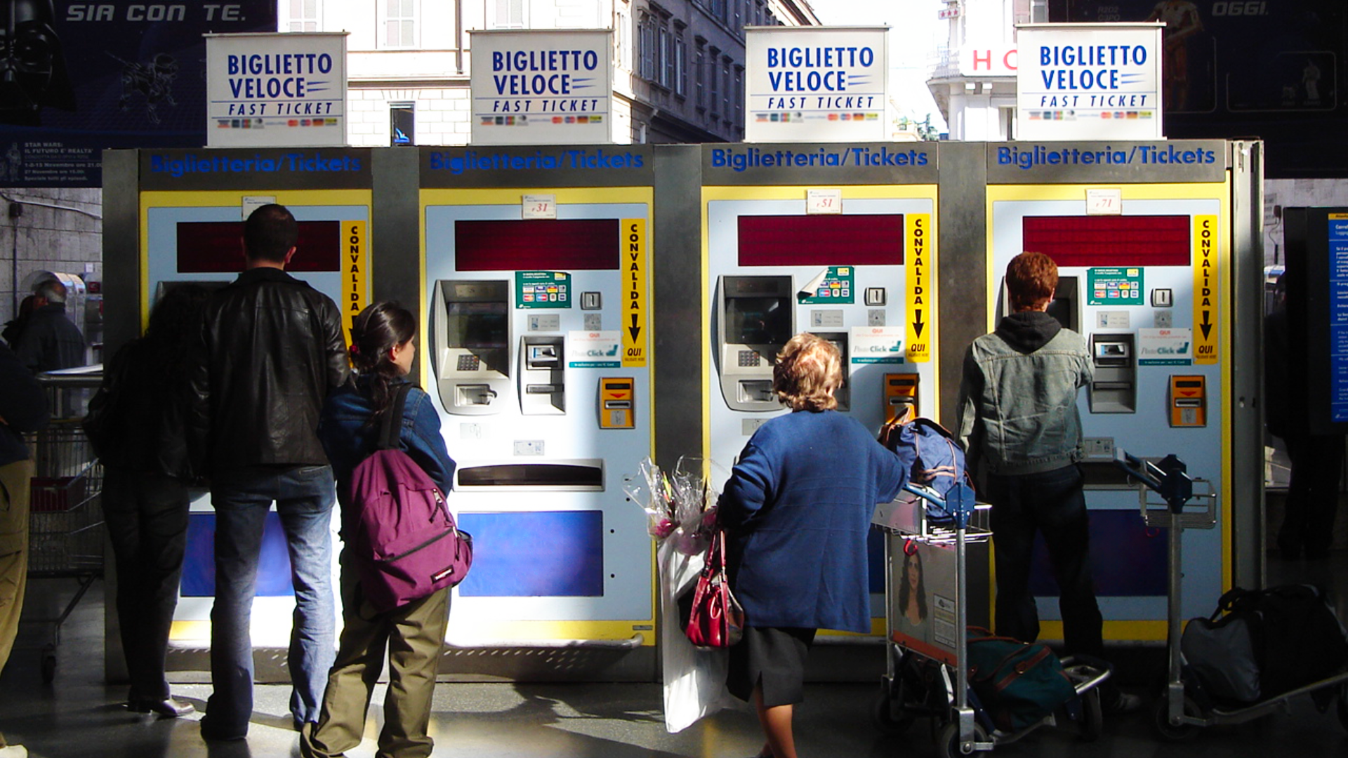 Ilustracja przedstawia cztery automaty samoobsługowe służące do zakupu biletów na pociąg. Przed automatami widoczni są ludzie: Po lewej wysoki mężczyzna kupuje bilet, obok niego stoi dziewczynka z plecakiem. Po prawej widoczna jest niska kobieta z bagażami oraz mężczyzna kupujący bilet w automacie. Ludzie stoją tyłem do kadru.