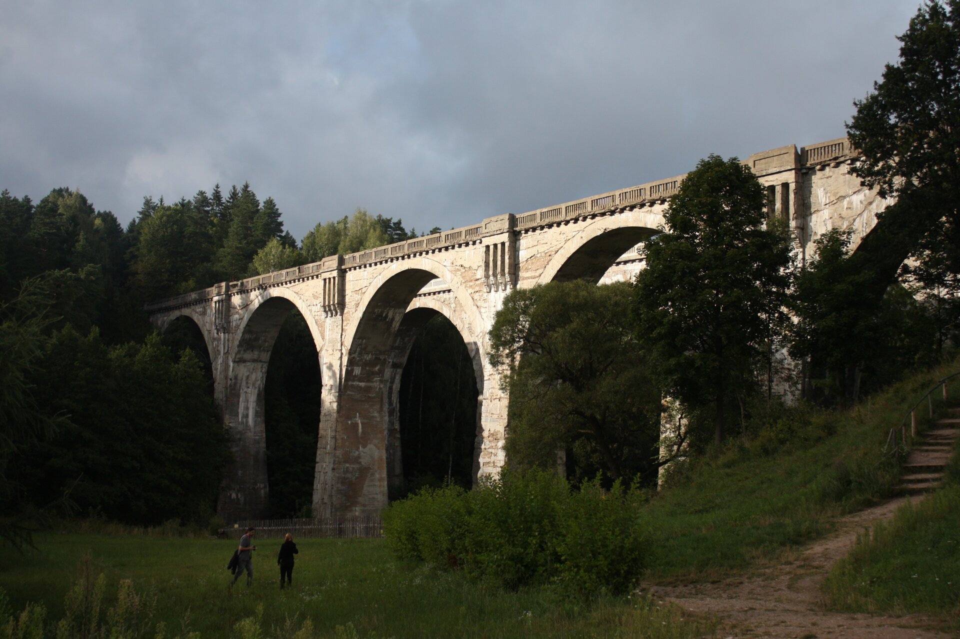 Na zdjęciu wysoki kamienny wiadukt kolejowy w pofałdowanym terenie. Dookoła drzewa. Pod wiaduktem ludzie.